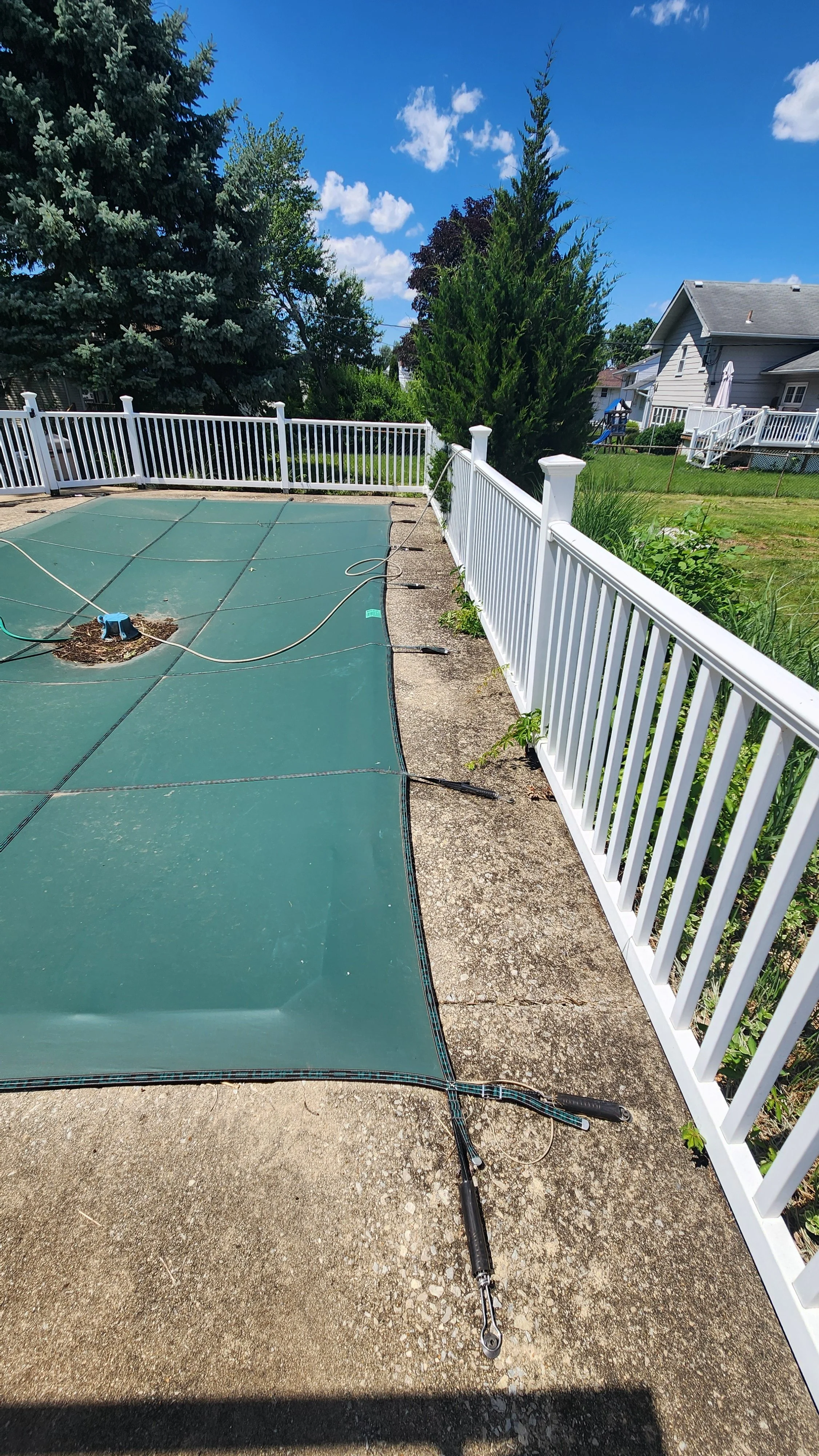 A backyard with a covered swimming pool with a green cover. The pool is surrounded by a white fence and is on a concrete patio. There are large trees and a blue sky with scattered clouds in the background before being cleaned.