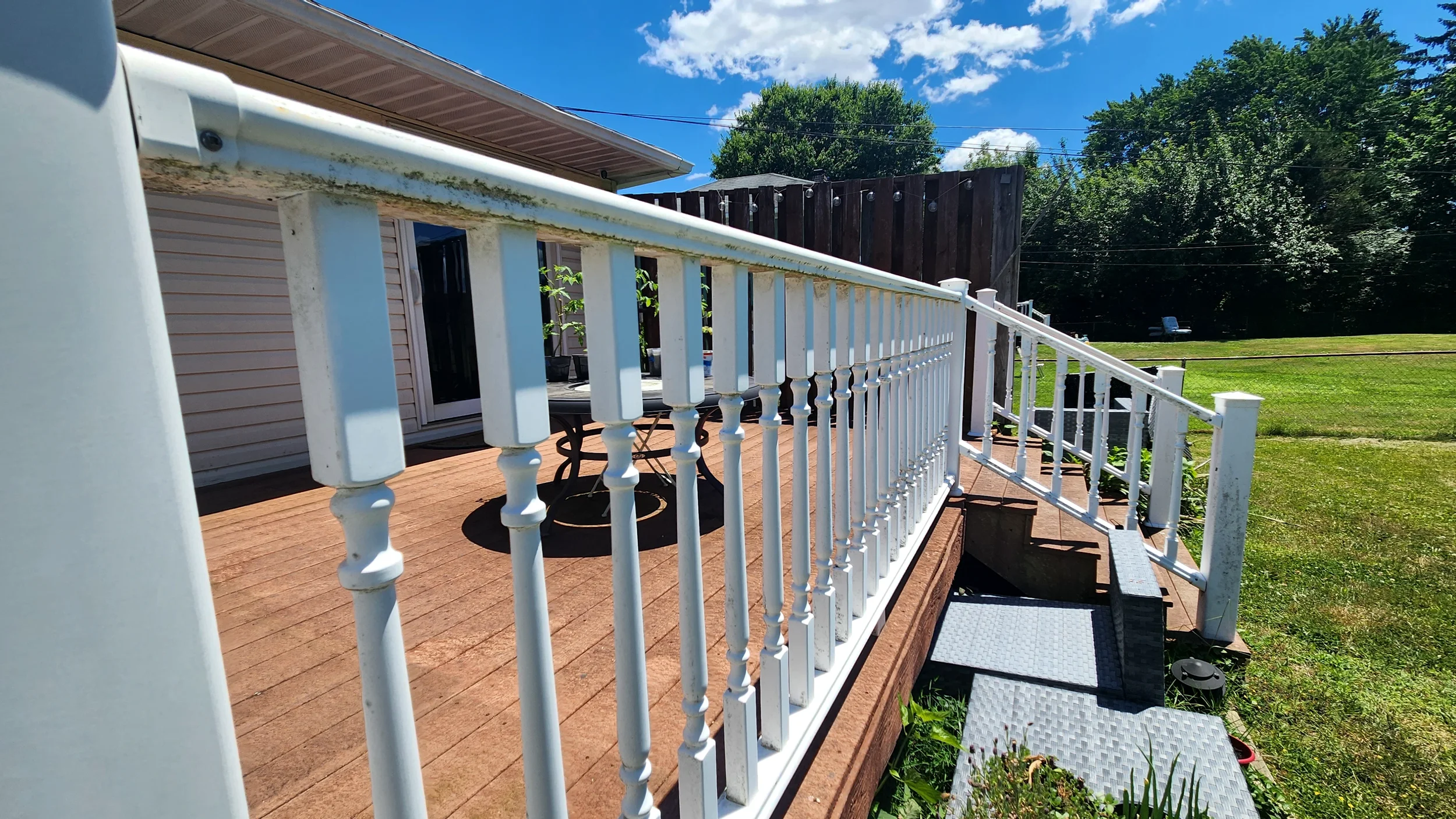 A wooden deck with a white metal railing leading down to a grassy yard, with trees and a blue sky with clouds in the background. Railings have algae covering them before being cleaned.