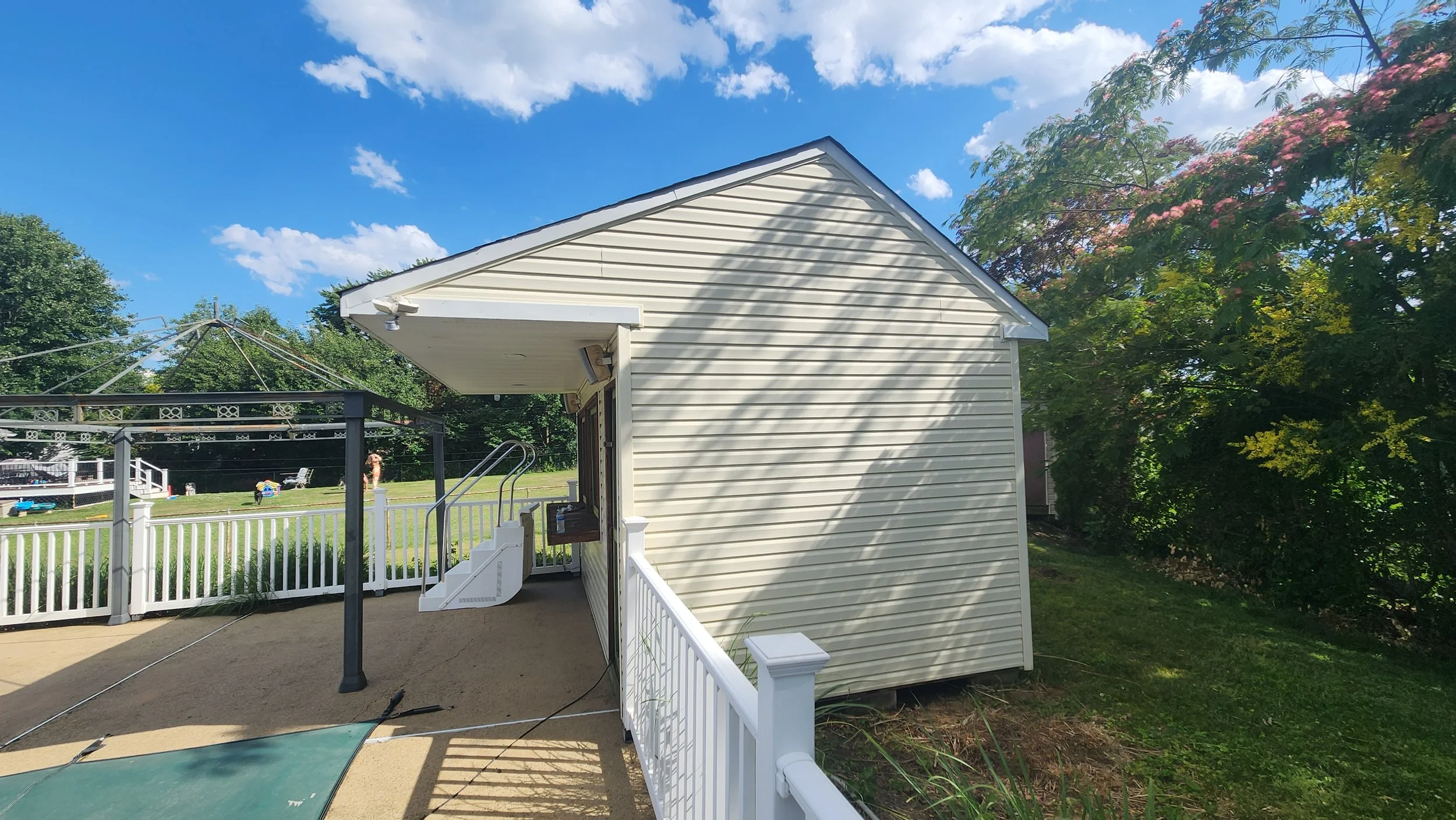 Backyard view with a white house and a fenced-in patio area, a grill, a clothes dryer, a tree with pink flowers, and a blue sky with clouds after the area has been cleaned.