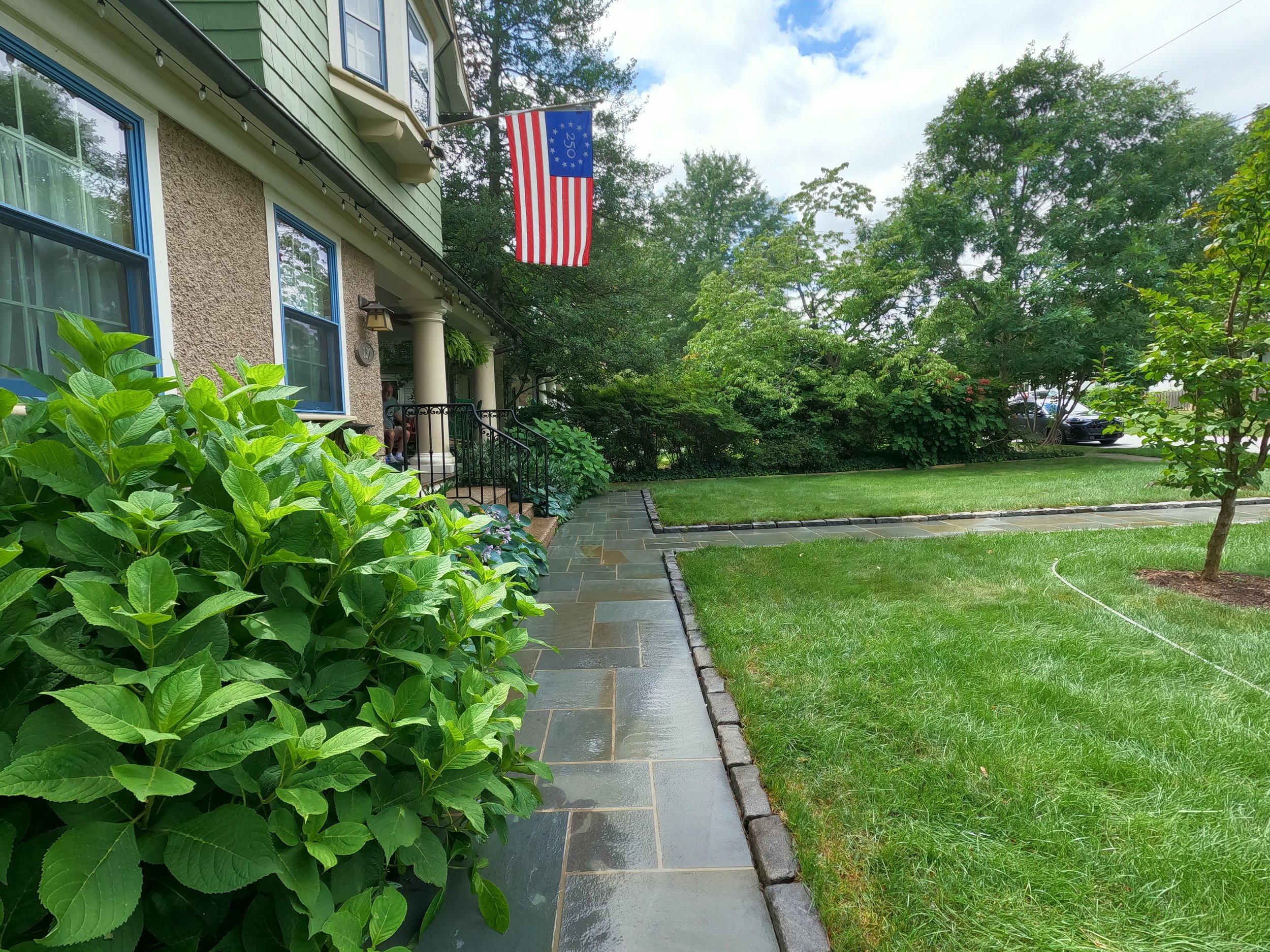 A front yard with a green lawn, a walkway with stone tiles, lush green bushes and trees, a house with green and beige exterior, blue window frames, porch with columns, and an American flag hanging from the house. This is after the walkway is cleaned.