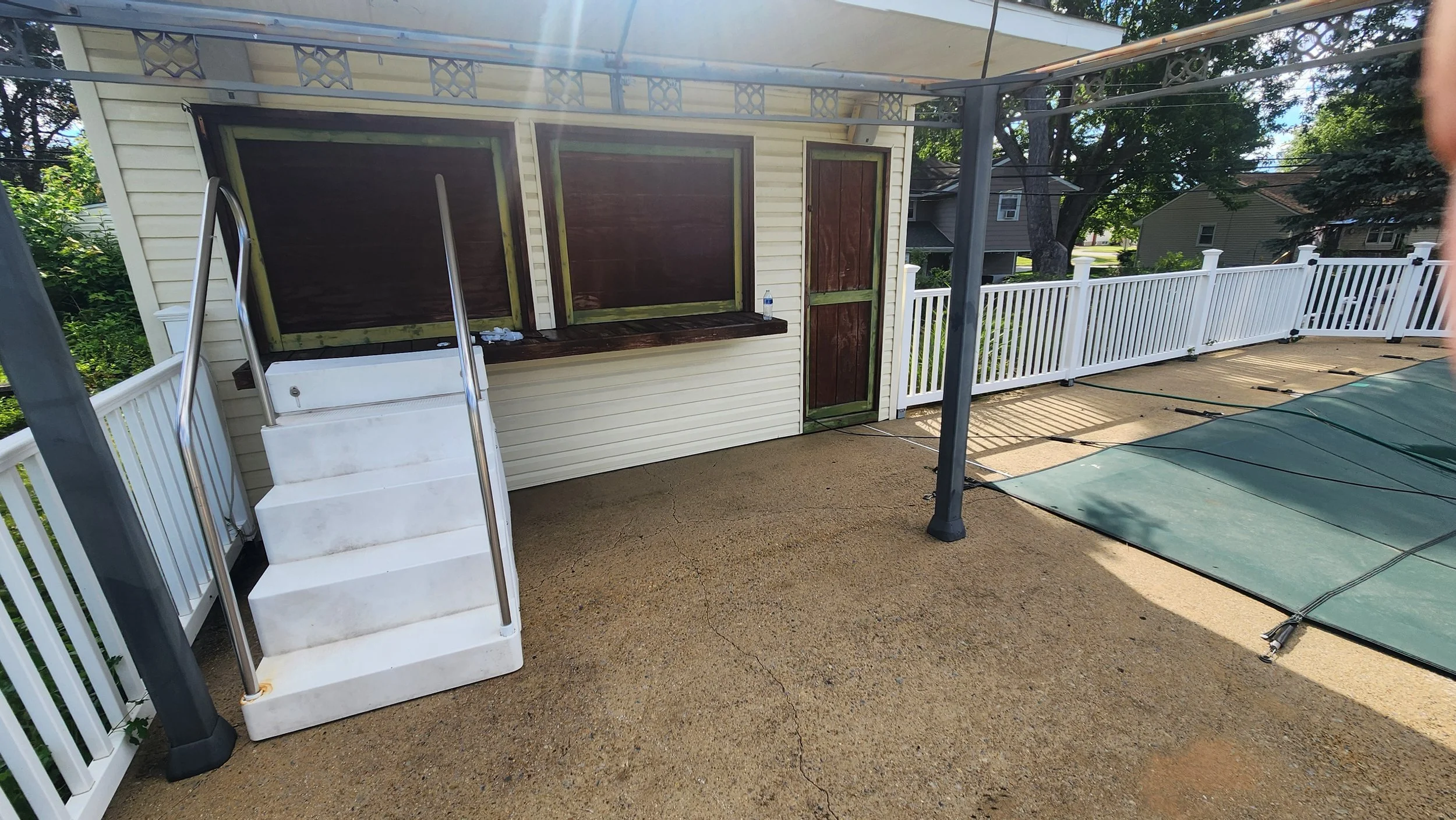 A backyard patio area featuring a small building with closed windows and a door, a white fence, a deck with canopy structure, and a covered swimming pool with a green cover after being cleaned.