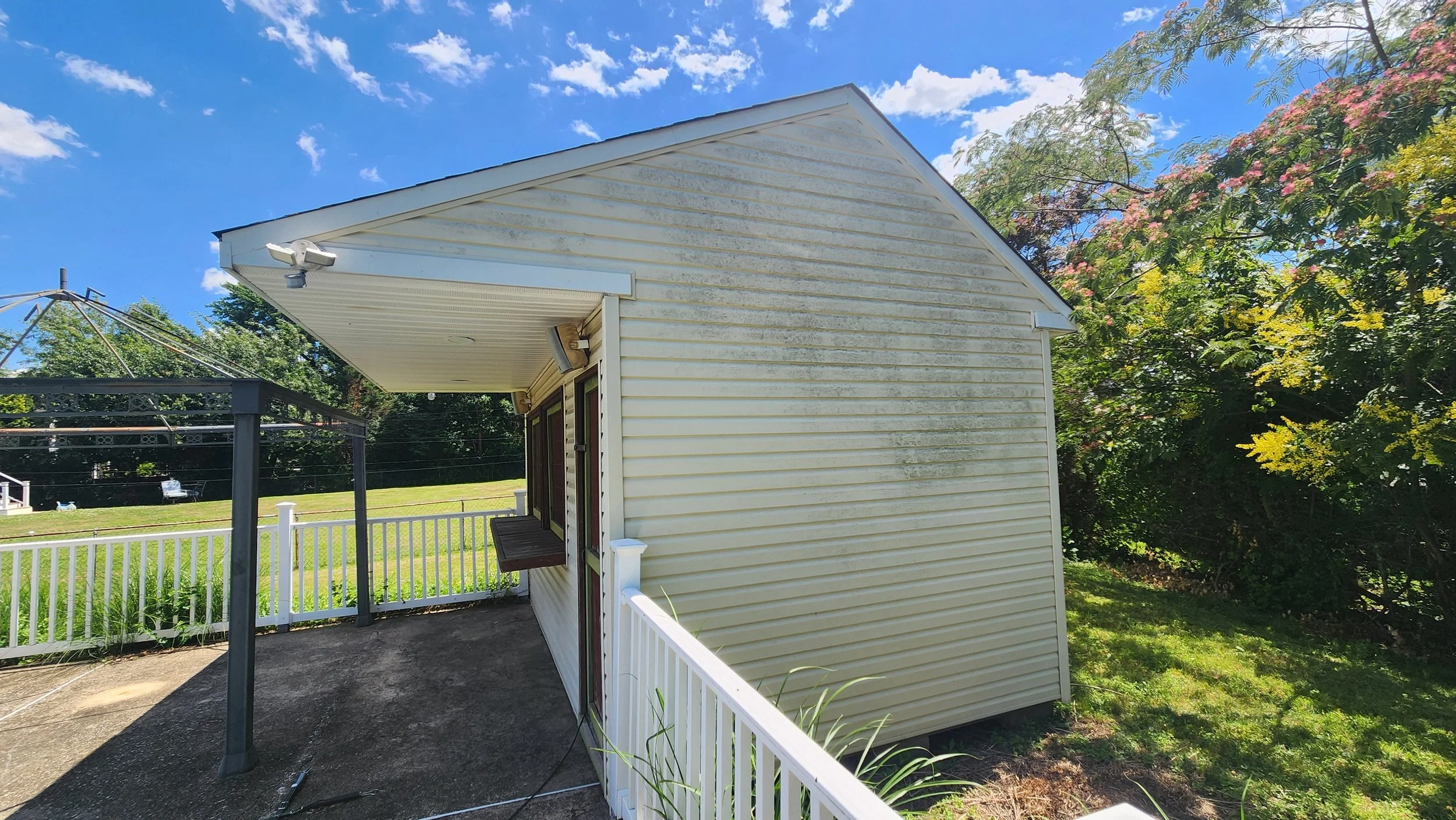 Backyard scene featuring a small white house with vinyl siding, a white fence, a grassy yard on the right, a paved area on the left, before being cleaned.