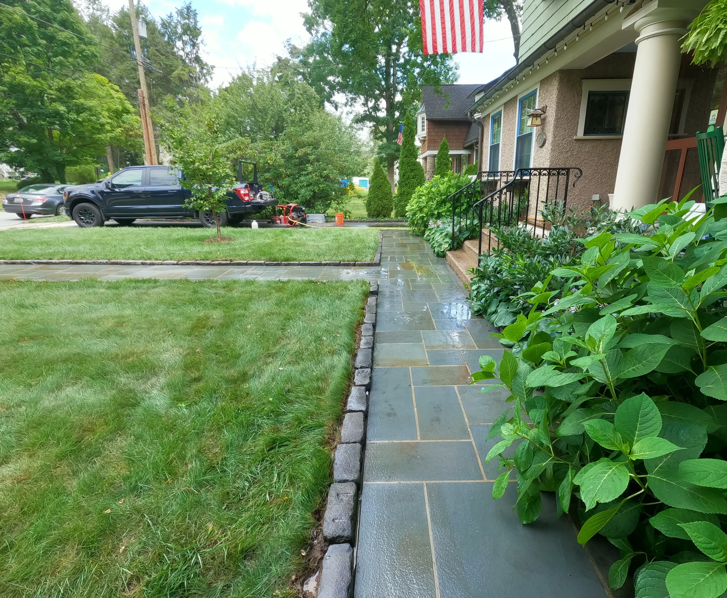 View of a residential front yard with green grass, a patio, and plants along the walkway. There is a house with beige stucco walls, white columns, blue window frames. This is after the walkway is cleaned.