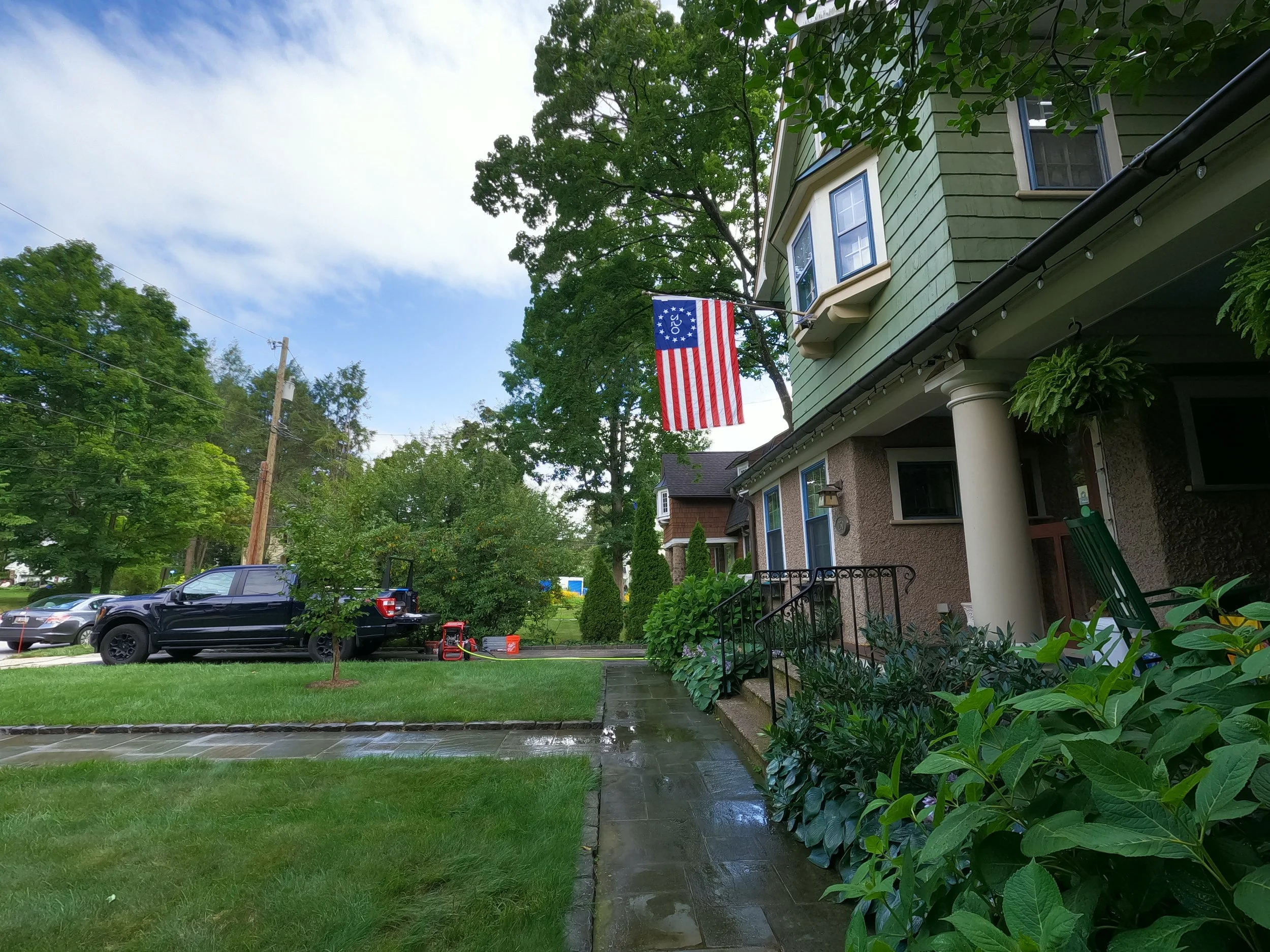 A suburban house with light green siding and white trim, with a front porch supported by a white column. A red, white, and blue American flag is hanging from a pole attached to the house. This is before the walkway is cleaned.