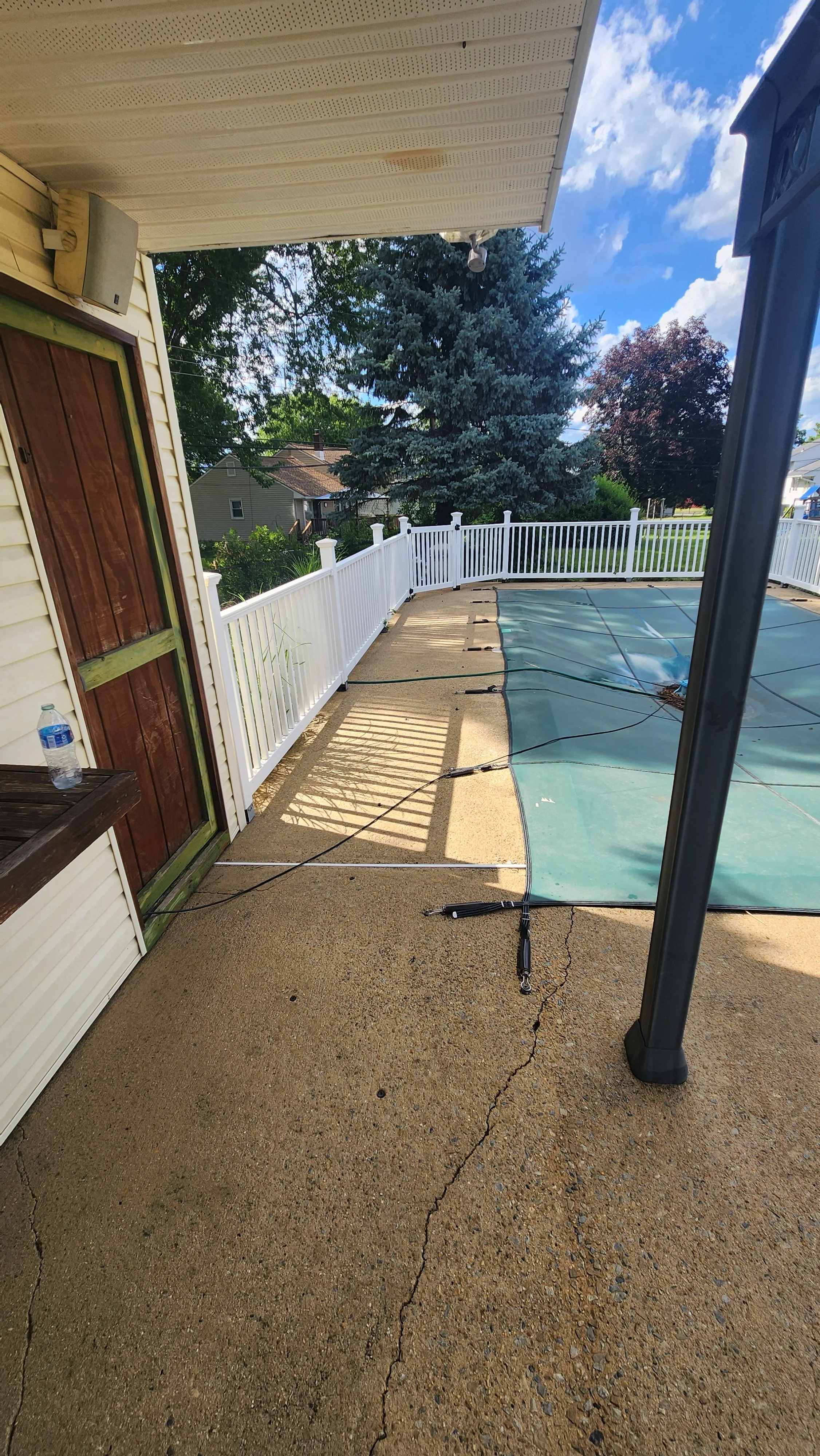 Backyard with a concrete patio, white vinyl fence, covered pool, large pine trees, and neighboring houses under a blue sky with clouds after being cleaned.