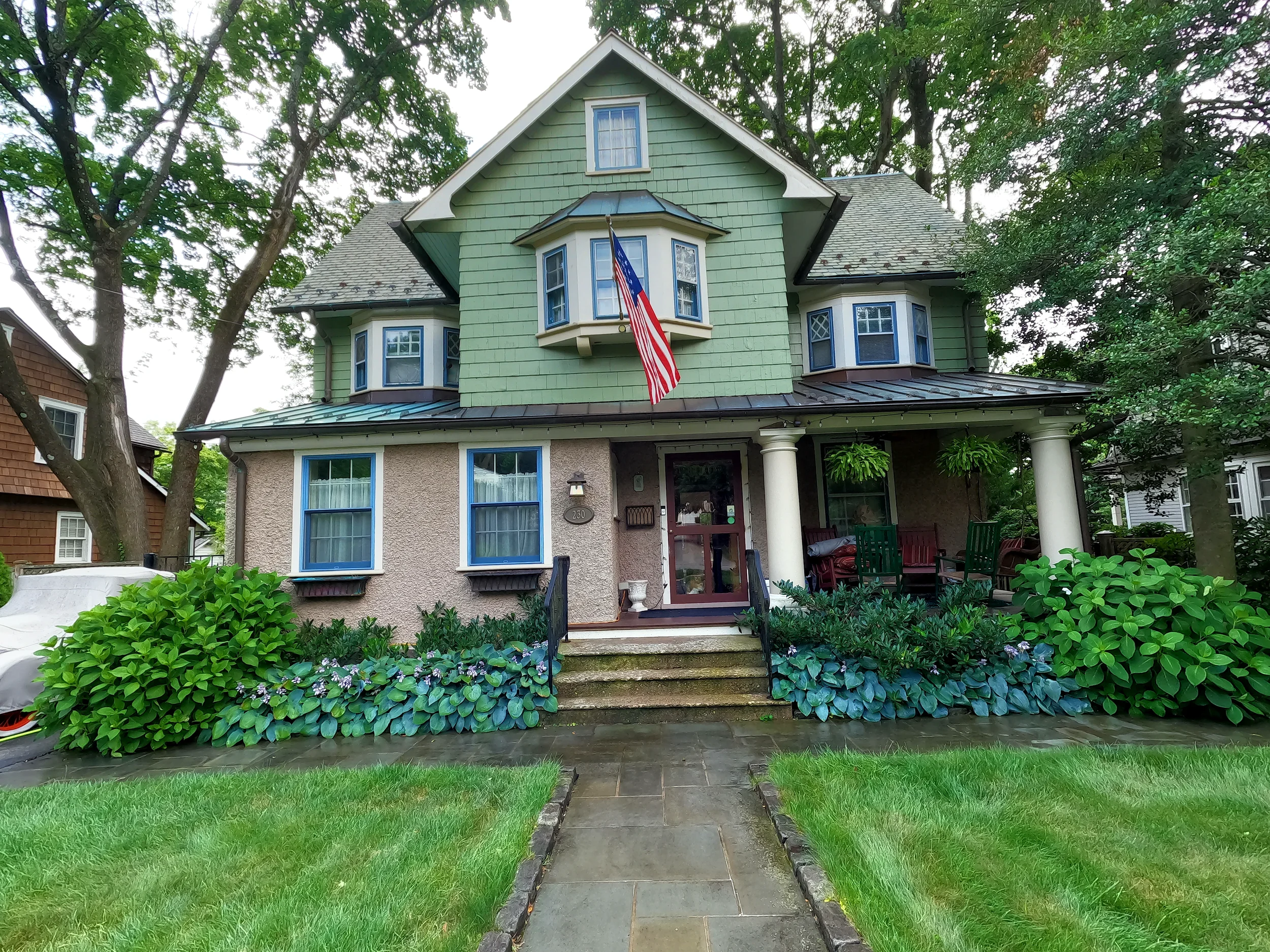 Front view of a green house with a variety of trees around it, American flag hanging from a central bay window, steps leading up to porch, and well-maintained garden with green bush and purple flowers in front. This is before cleaning the walkway.