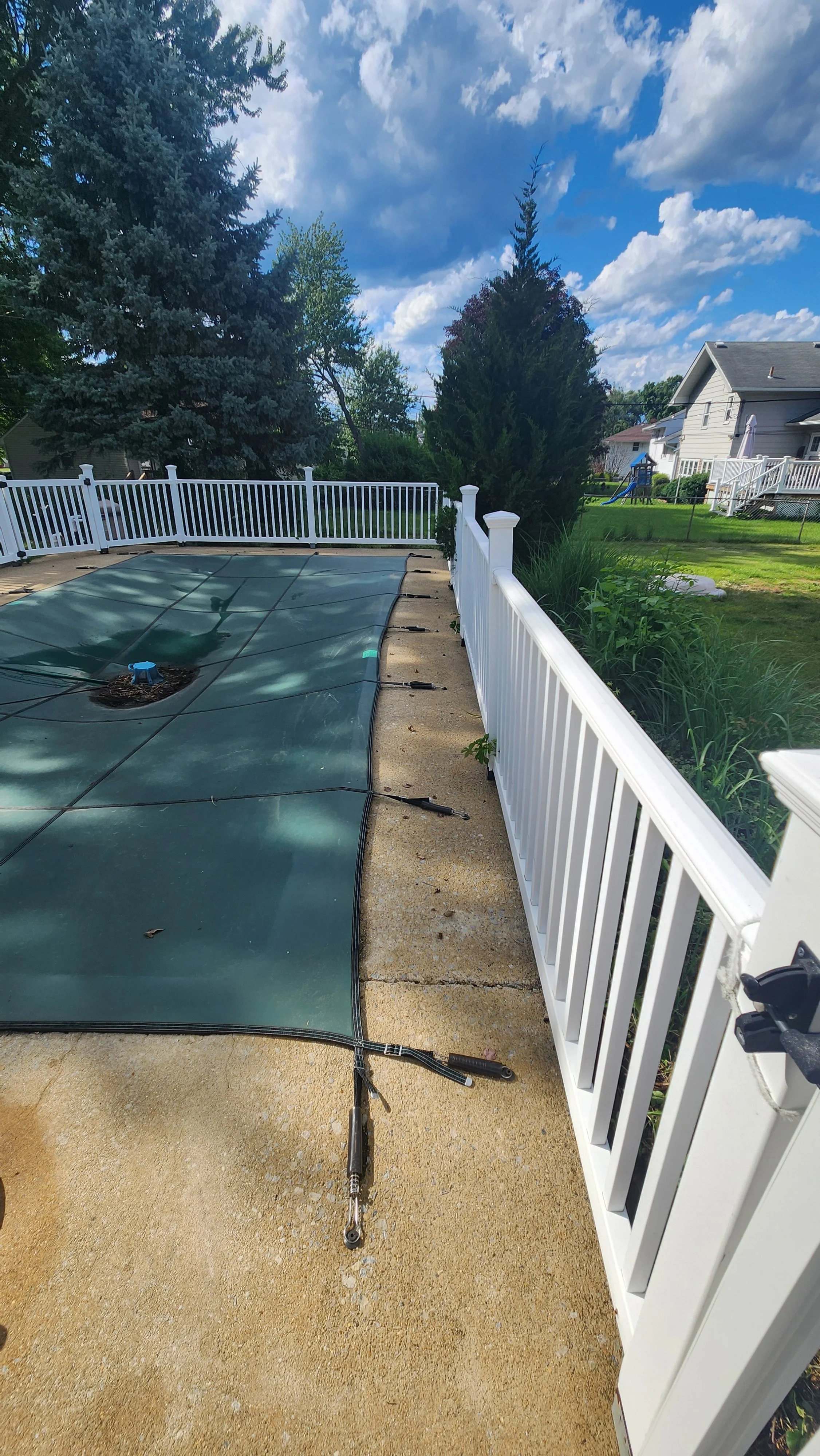 Backyard with a swimming pool covered with a green safety cover, surrounded by a concrete patio and a white vinyl fence. There are trees and houses in the background under a partly cloudy sky after being cleaned.