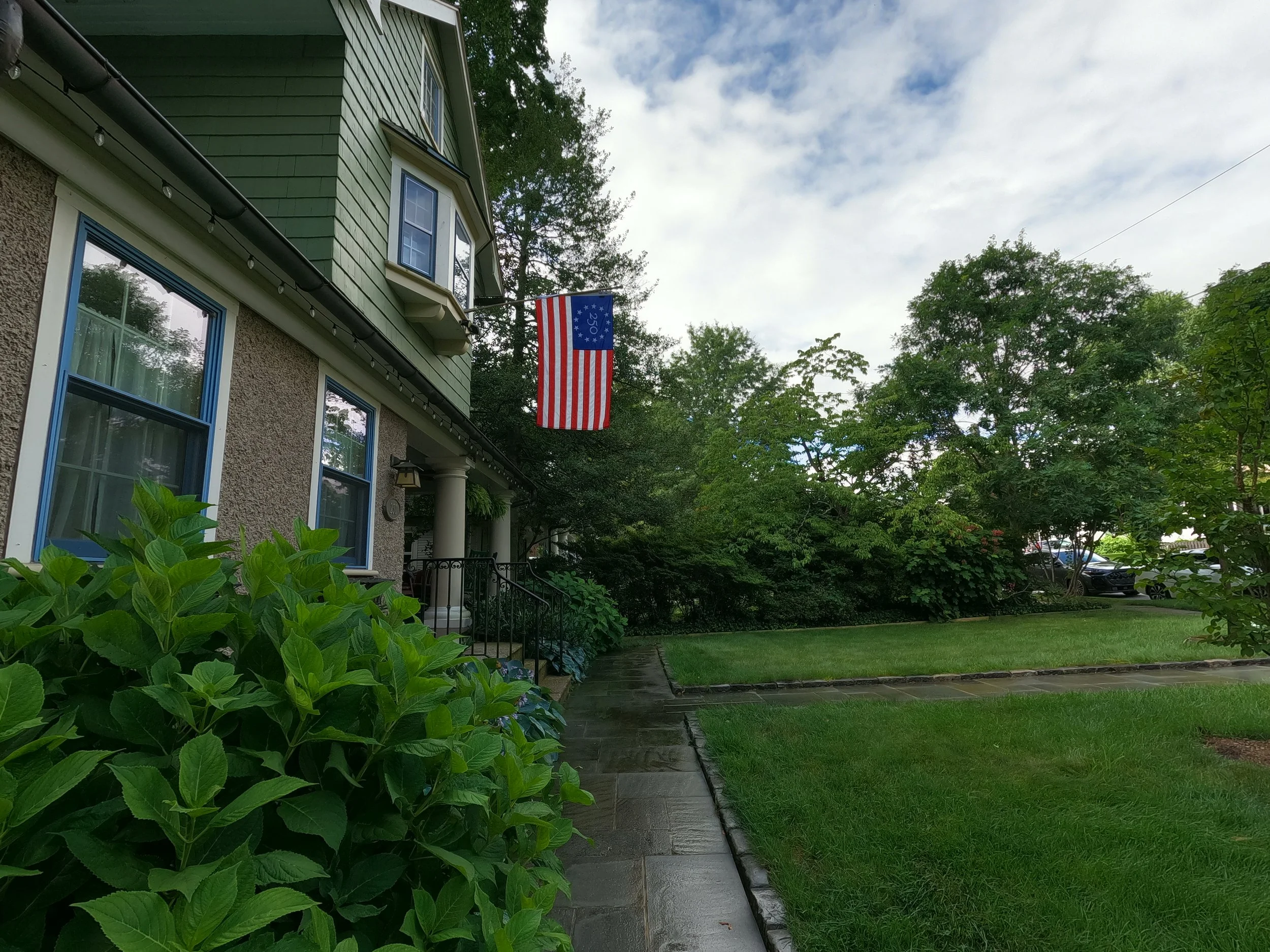A house with green siding and beige textured stucco, decorated with string lights, has a porch with white columns and a small American flag hanging from the roof. This is before the walkway is cleaned.