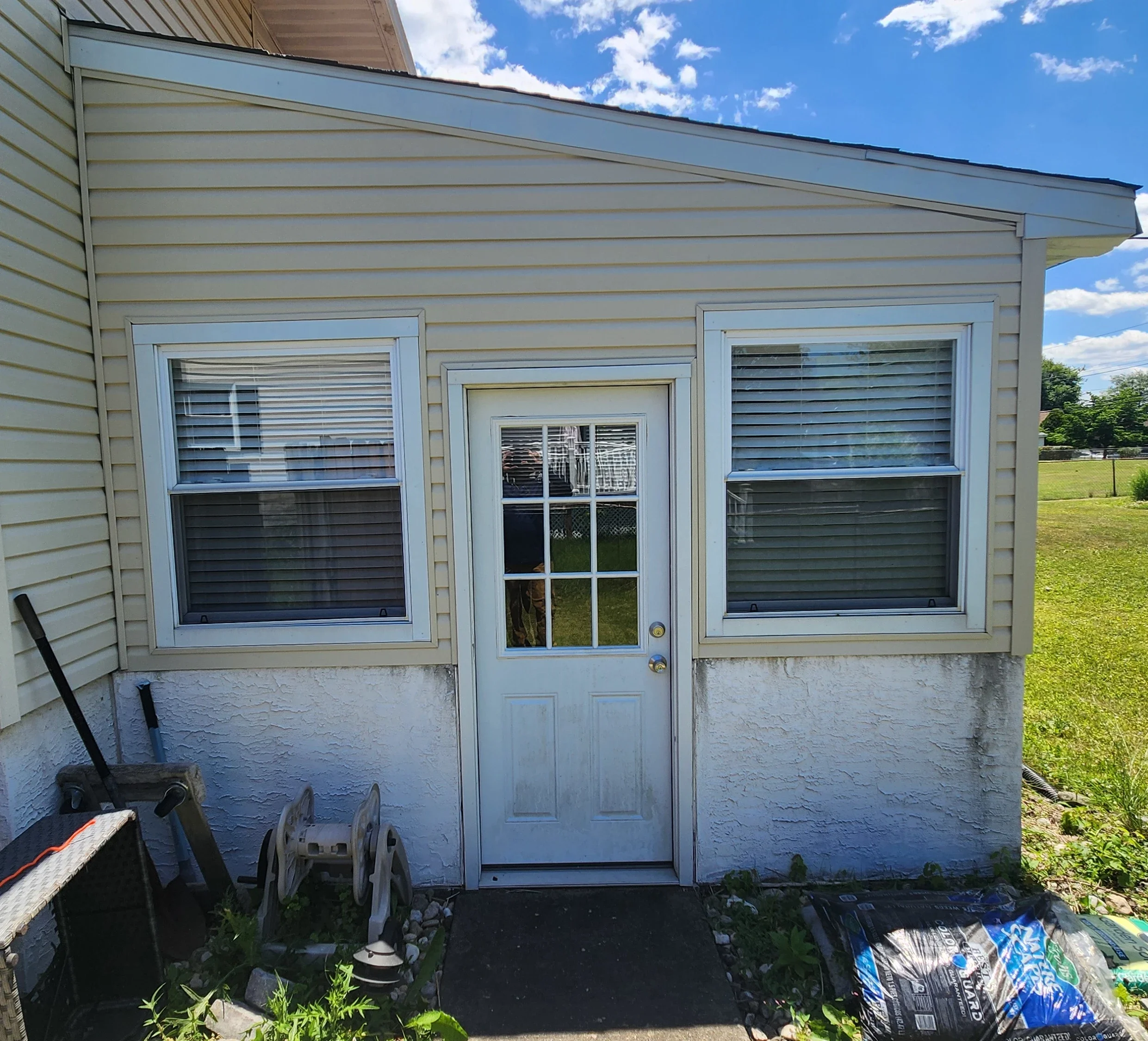 Backyard scene showing a section of a house with beige siding, two windows, and a door. There are gardening chairs, a bag of soil or mulch, and some garden tools on the ground, with grass and trees visible in the background. Before being cleaned.
