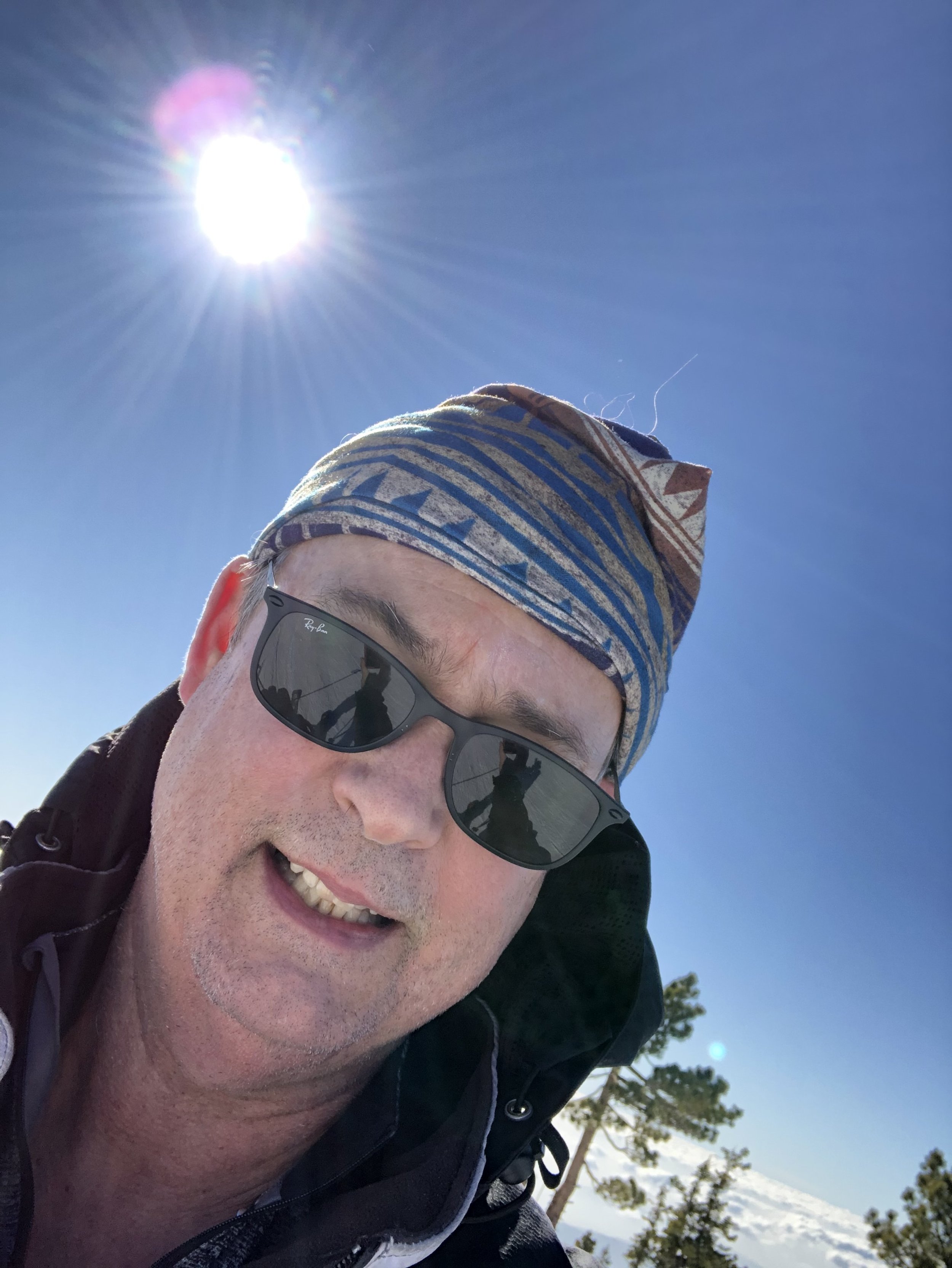 Man wearing sunglasses and a patterned bandana, smiling under a bright sun with a clear blue sky and trees in the background.