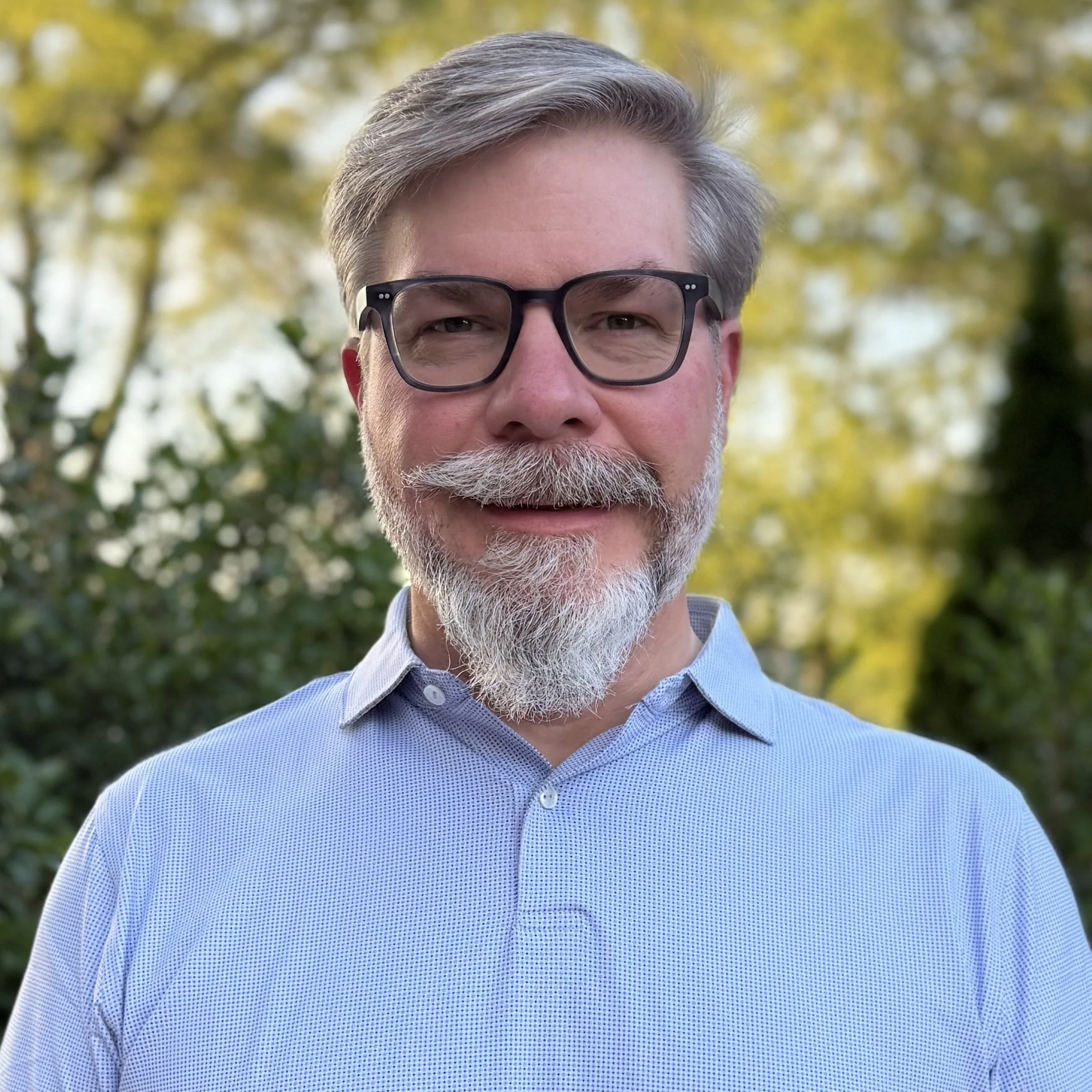 A middle-aged man with gray hair, glasses, and a beard, wearing a light blue dress shirt, standing outdoors with a background of green and yellow trees.