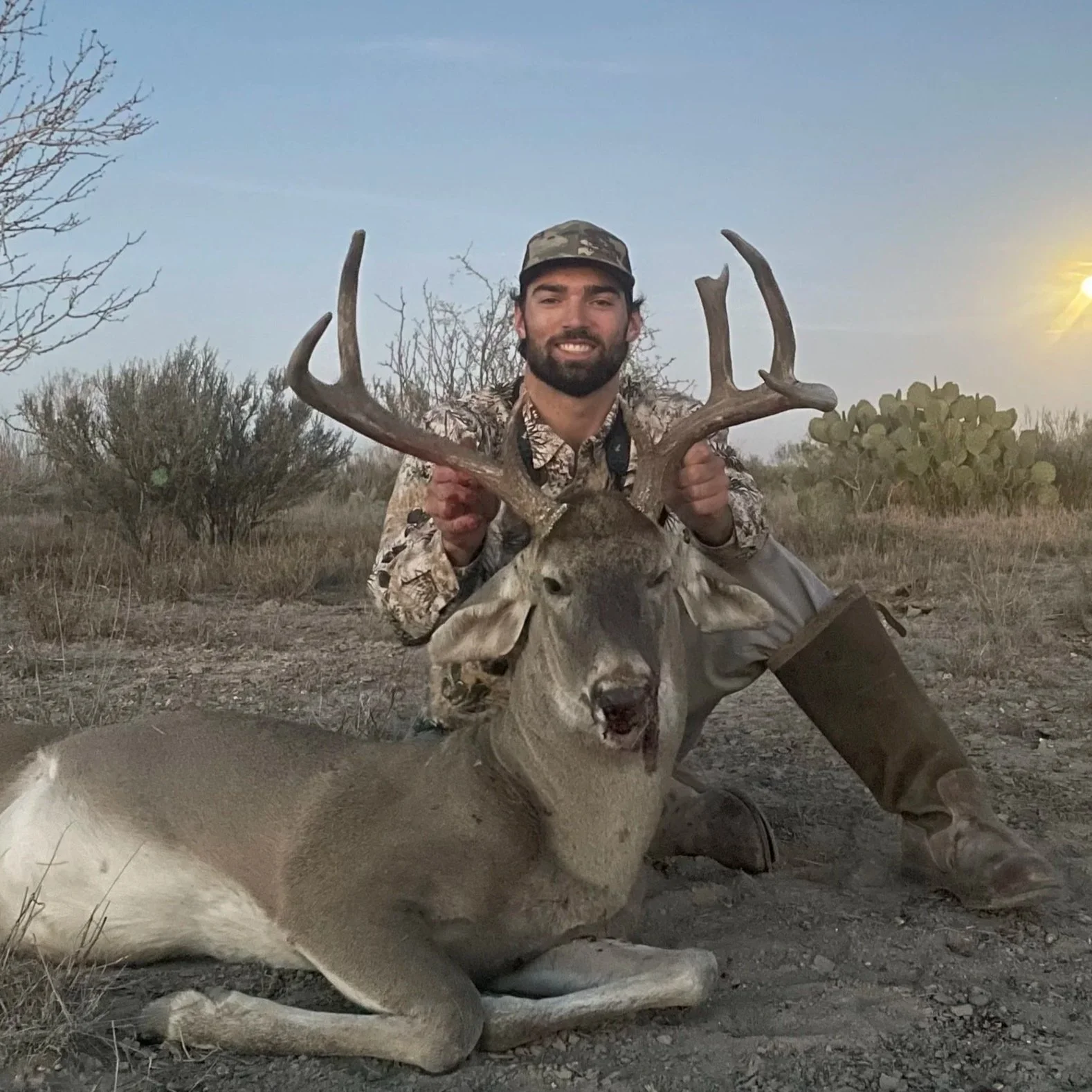 A man in camouflage clothing kneels next to a large deer with antlers, holding its antlers, in a desert landscape at sunset.