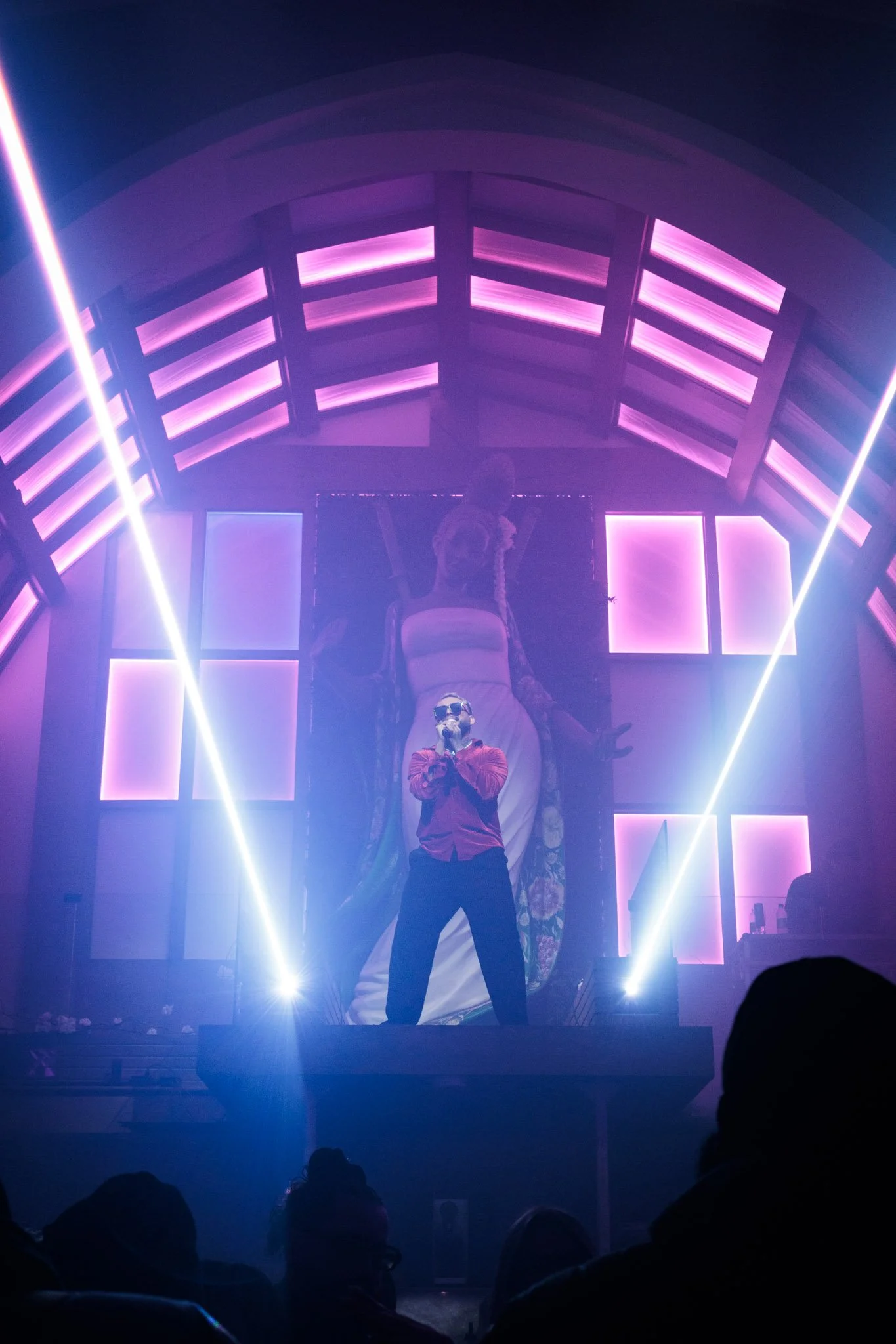 A person wearing sunglasses and a red shirt performing on stage with purple and pink lighting, a large screen with an artistic image of a woman in the background, and audience silhouettes in the foreground.