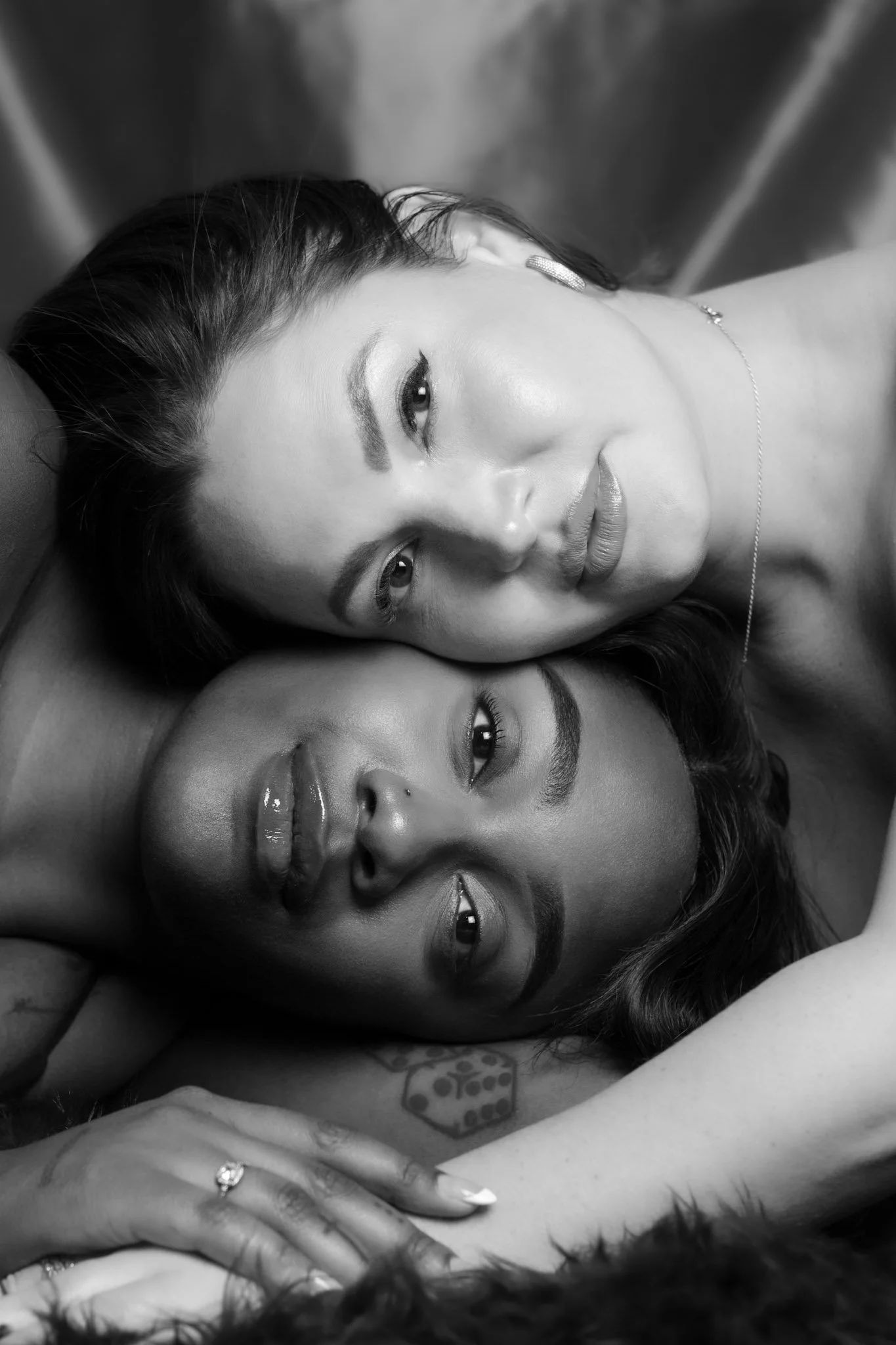 Two women lying close together, one with light skin and the other with dark skin, both with dark hair, in a black-and-white photograph. They are resting their heads on each other and looking at the camera.