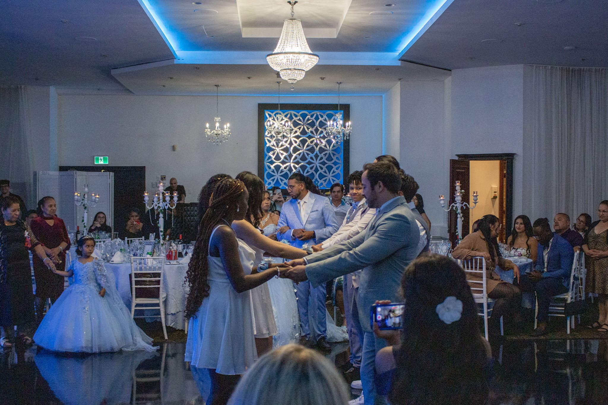 A wedding reception inside a banquet hall with a bride and groom holding hands in the center, surrounded by guests seated at decorated tables, with chandeliers hanging from the ceiling and a decorative wall in the background.