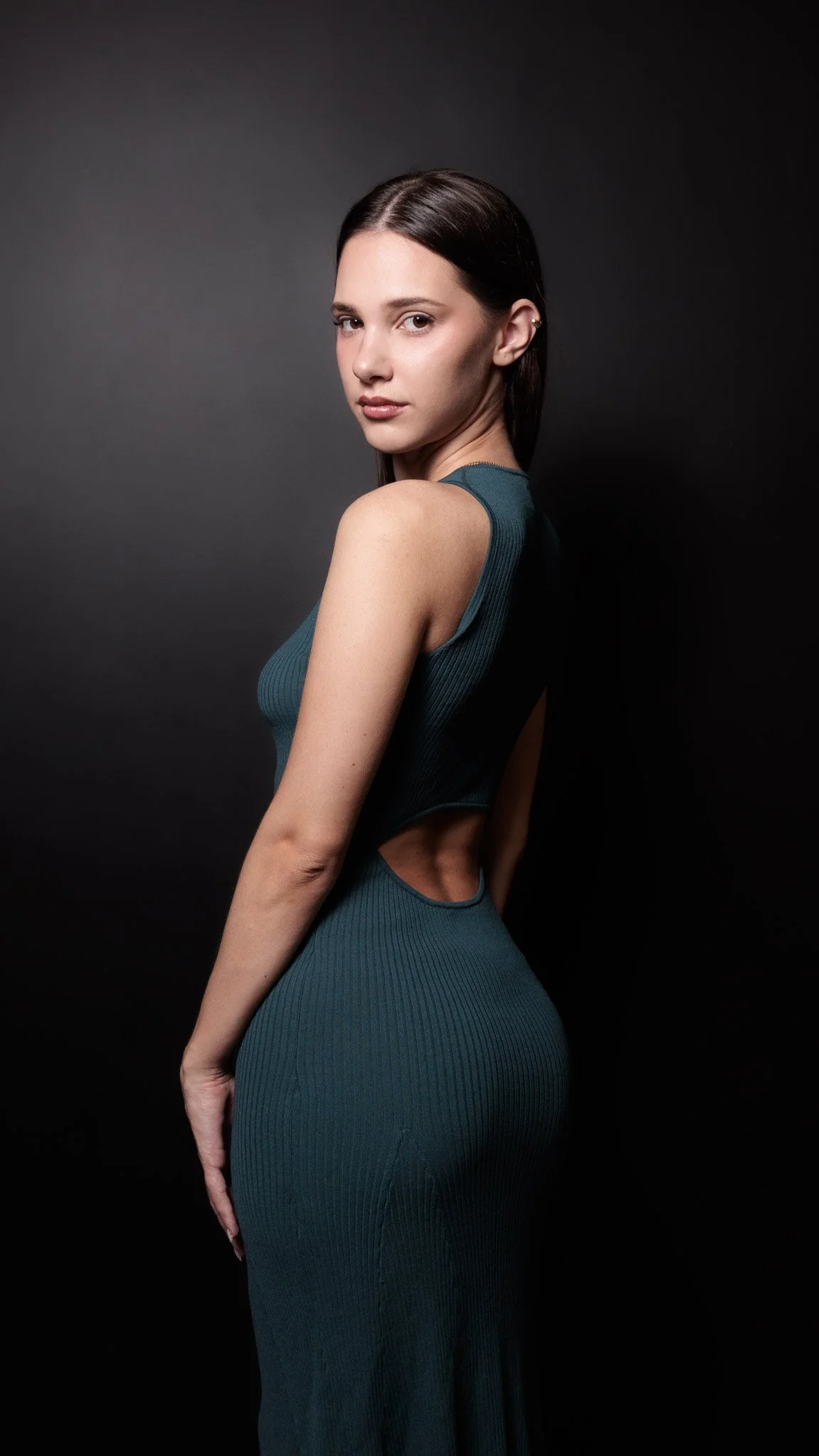 A young woman with dark hair styled straight, wearing a sleeveless teal ribbed dress with cutouts at the waist, posing against a dark background.