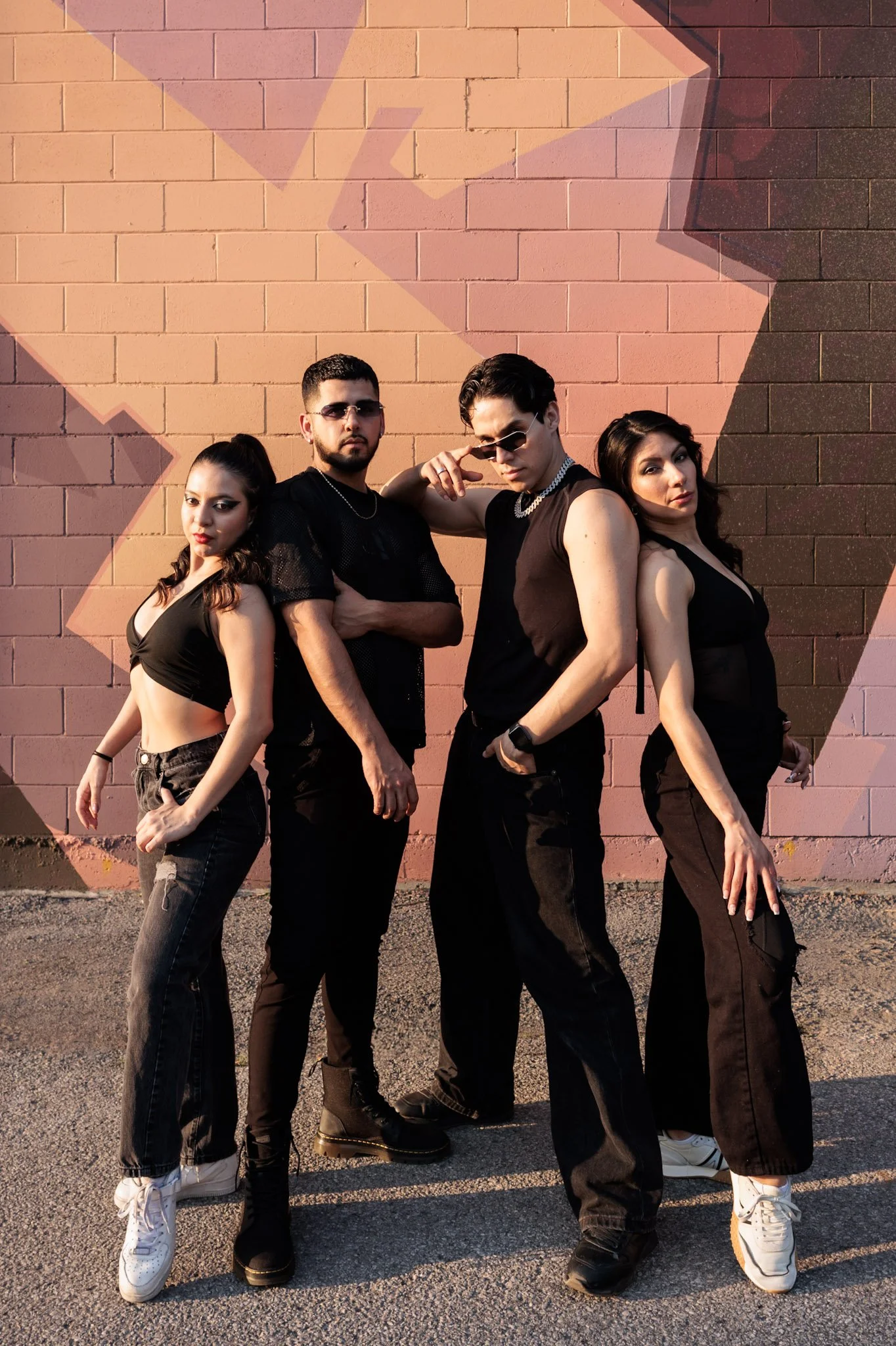 Four young adults standing against a pink graffiti wall, dressed in black casual streetwear, posing confidently.