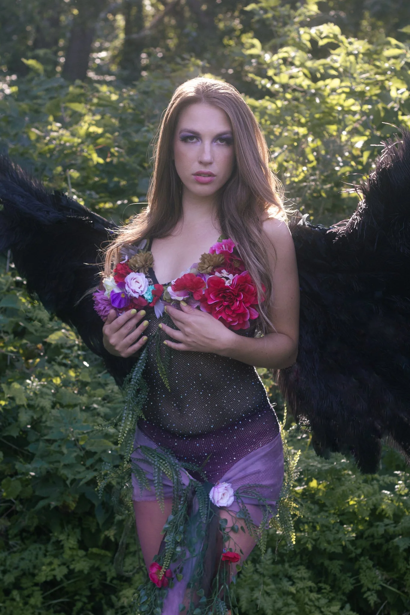 A woman with long brown hair stands in a forest, holding flowers over her chest and wearing black feathered wings. She is dressed in a sheer, beaded top and a pink, semi-transparent skirt decorated with flowers and greenery.