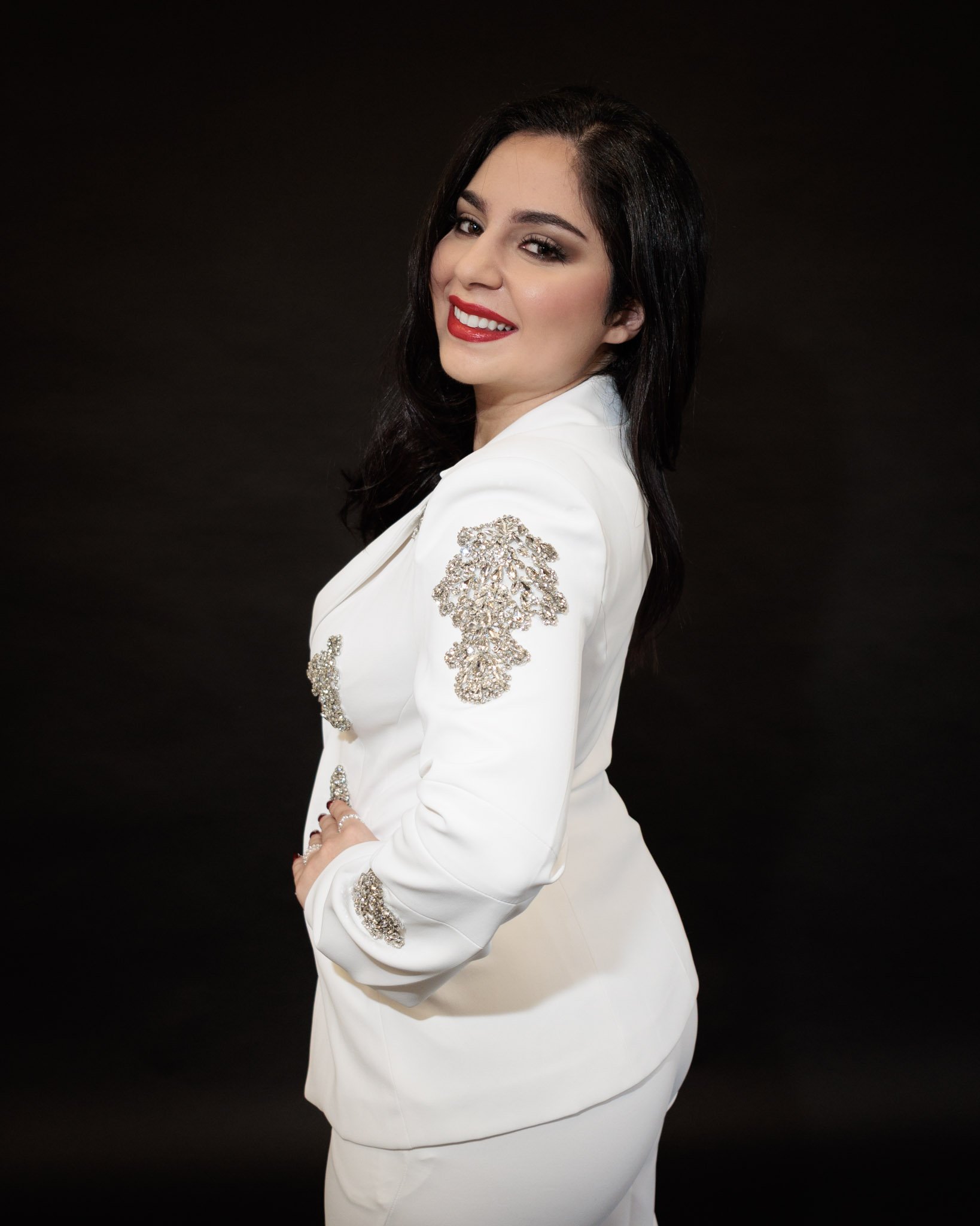 A woman with dark hair and red lipstick wears a white suit with silver embellishments, posing against a black background.