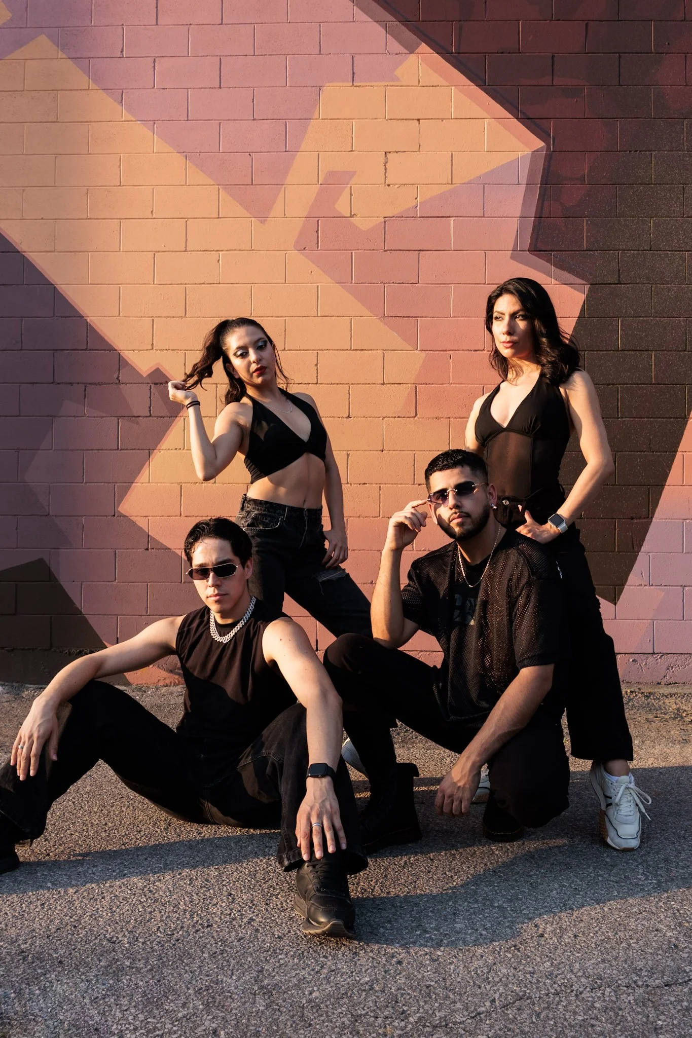 Four young adults posing outdoors in front of a colorful abstract wall mural, dressed in black clothing with sunglasses, striking confident poses during sunset.