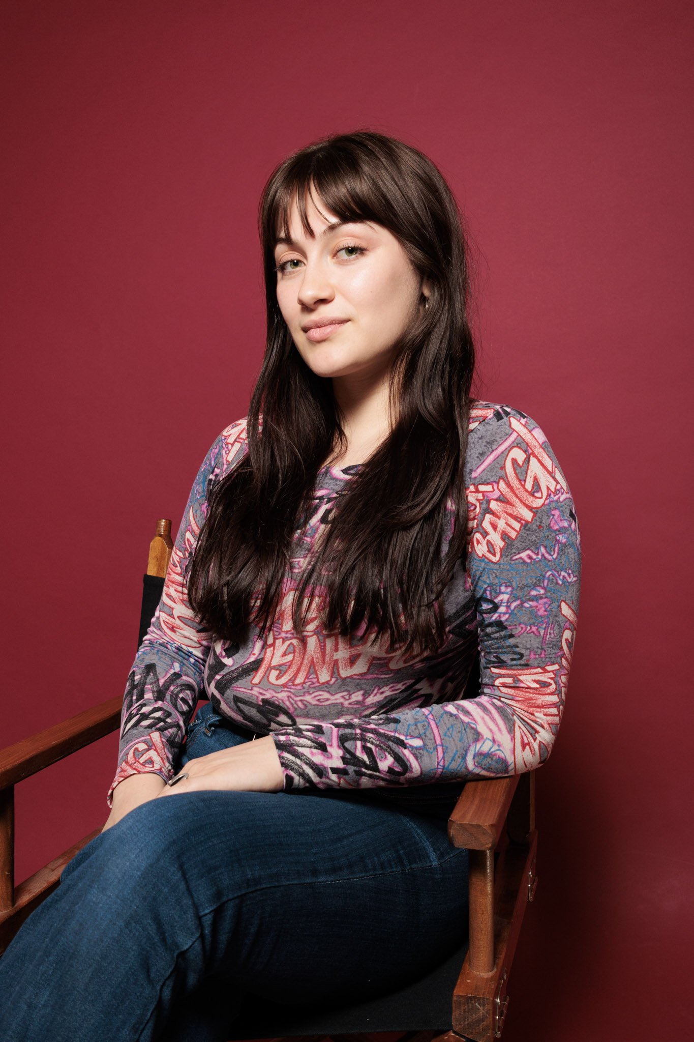 Young woman with long brown hair sitting on a wooden director's chair against a pink background, wearing a colorful graphic long-sleeve shirt and blue jeans.