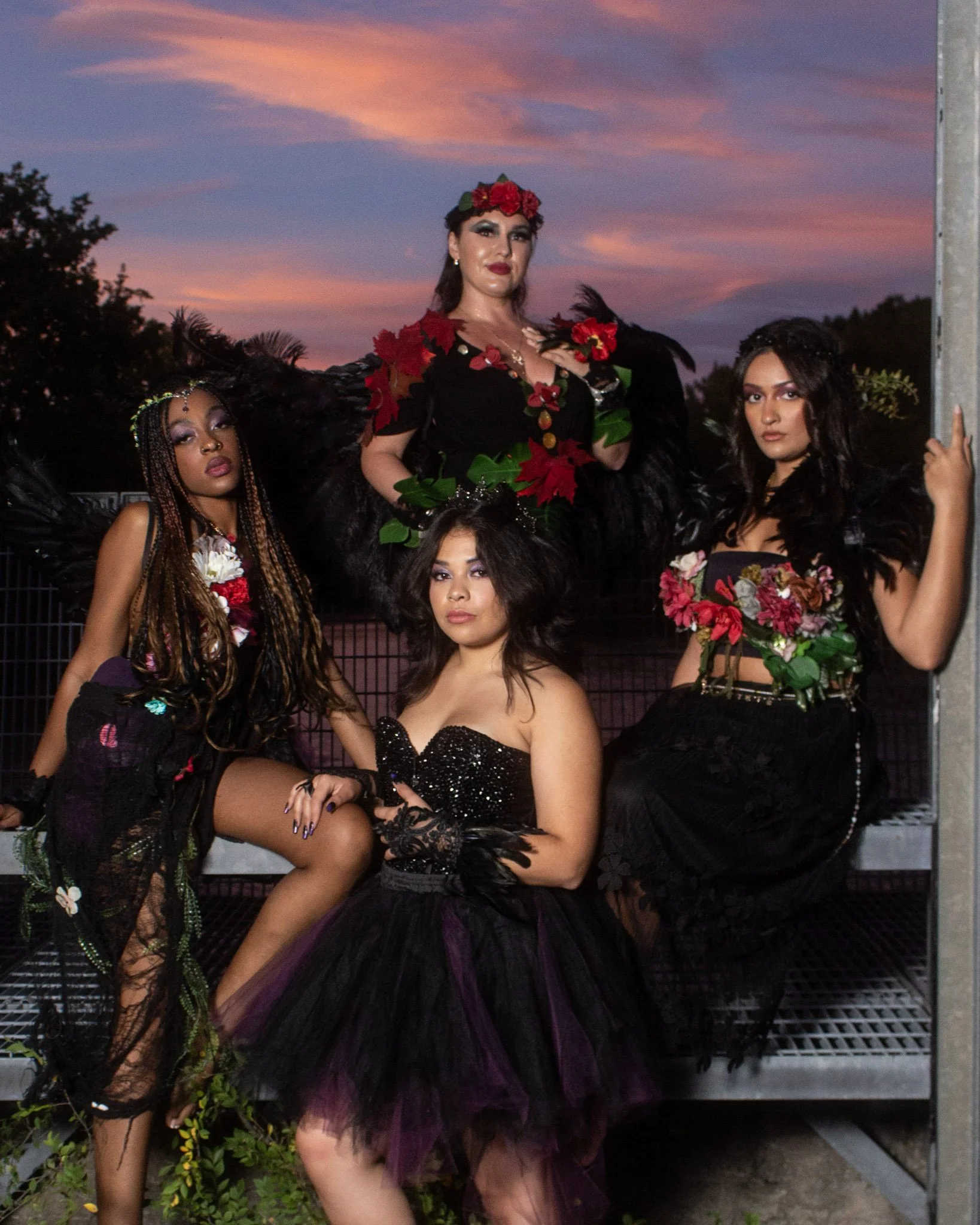 Four women dressed in black and decorated with flowers, posed outdoors during sunset with colorful sky in the background.