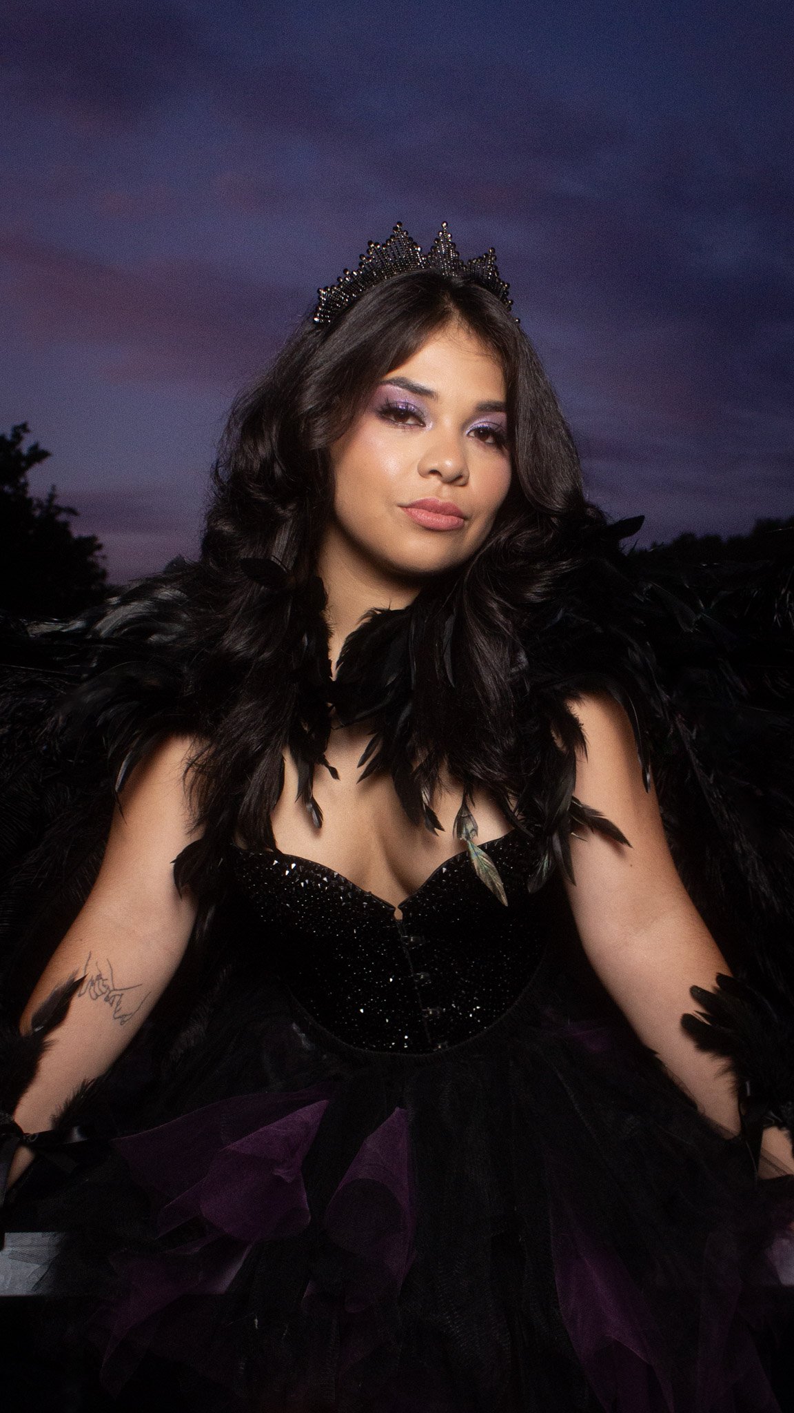 A woman dressed as a dark fairy or night queen, wearing a black bejeweled corset, black feathered wings, and a crown, with a sunset or dusk sky in the background.