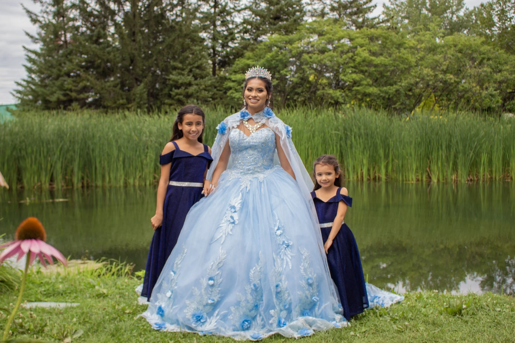 A woman dressed as a princess in a light blue gown with blue floral accents and a silver crown poses outdoors with two young girls wearing matching dark blue dresses with off-the-shoulder sleeves near a pond with greenery and trees in the background.