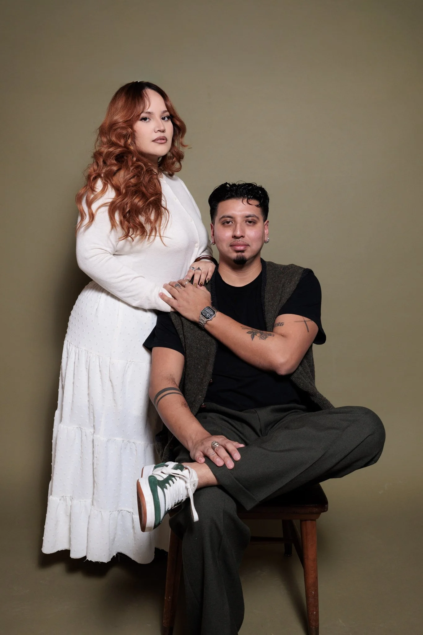 A man sitting on a chair with a woman standing beside him, both looking at the camera against a plain background.