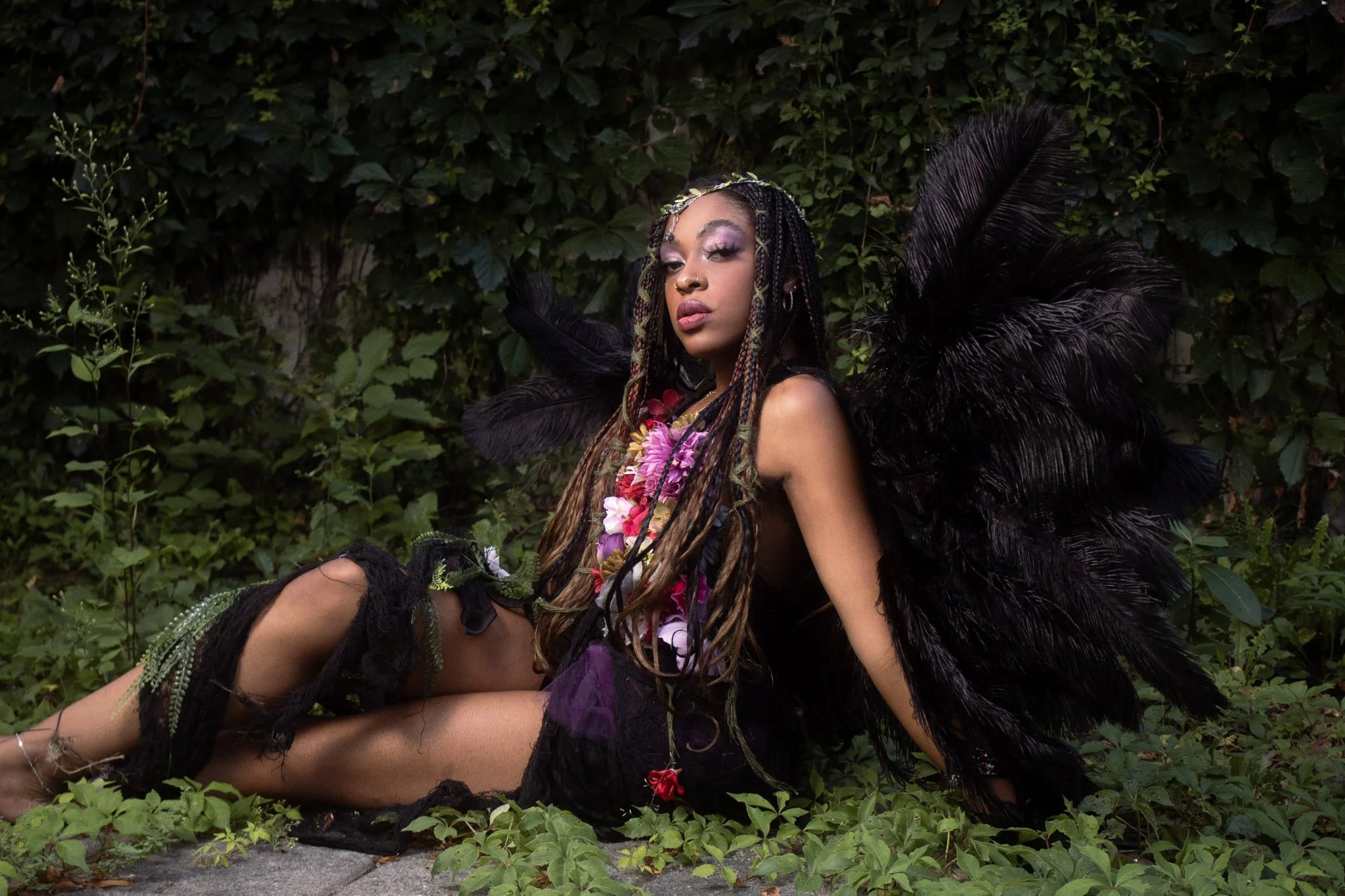 A woman dressed as a dark fairy or mythical creature with black feathers on her shoulders, floral adornments on her chest, and long, braided hair, lying on the ground amid green foliage.