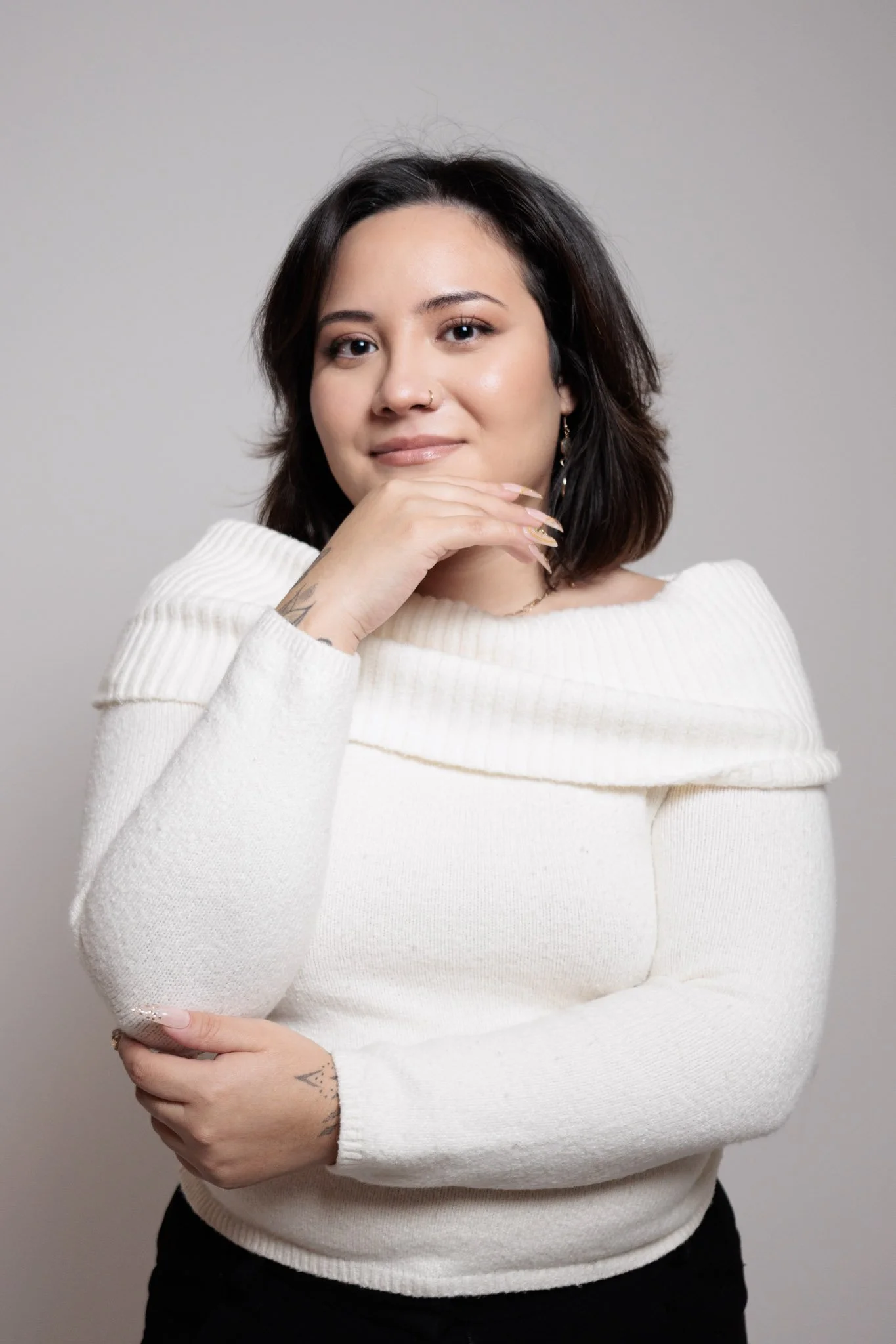 A young woman with dark hair and tattoos, wearing a white sweater, is posing with her left arm crossed and her right hand resting near her chin, against a neutral background.