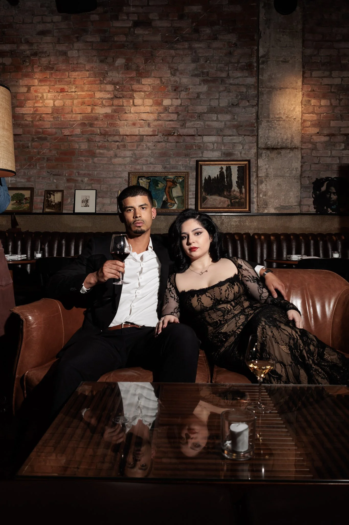 A man and woman sitting on a leather couch in a dimly lit, stylish room with a brick wall, art on the wall, and a glass of red wine and white wine on a wooden table in front of them. Roseline Montréal. 