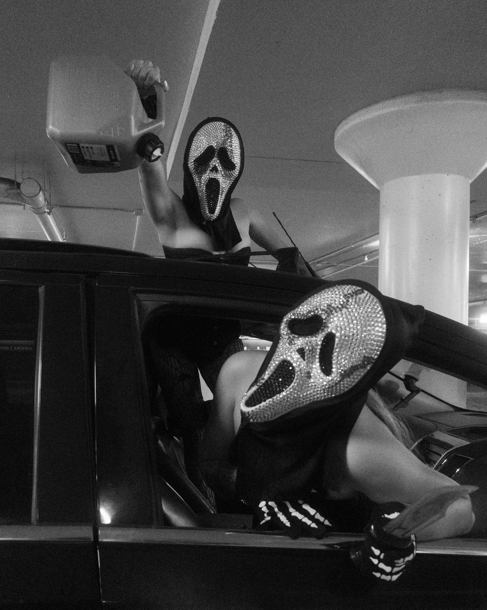 Black and white photo of two people inside a car, both wearing masks with a screaming face design and rhinestones. One person is on the roof holding a container, and the other is leaning out of the car window.