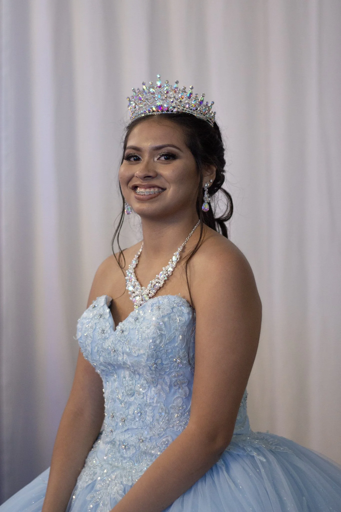 A young woman wearing a light blue, embroidered quinceañera dress with a tulle skirt, a tiara, and jewelry, smiling at the camera.