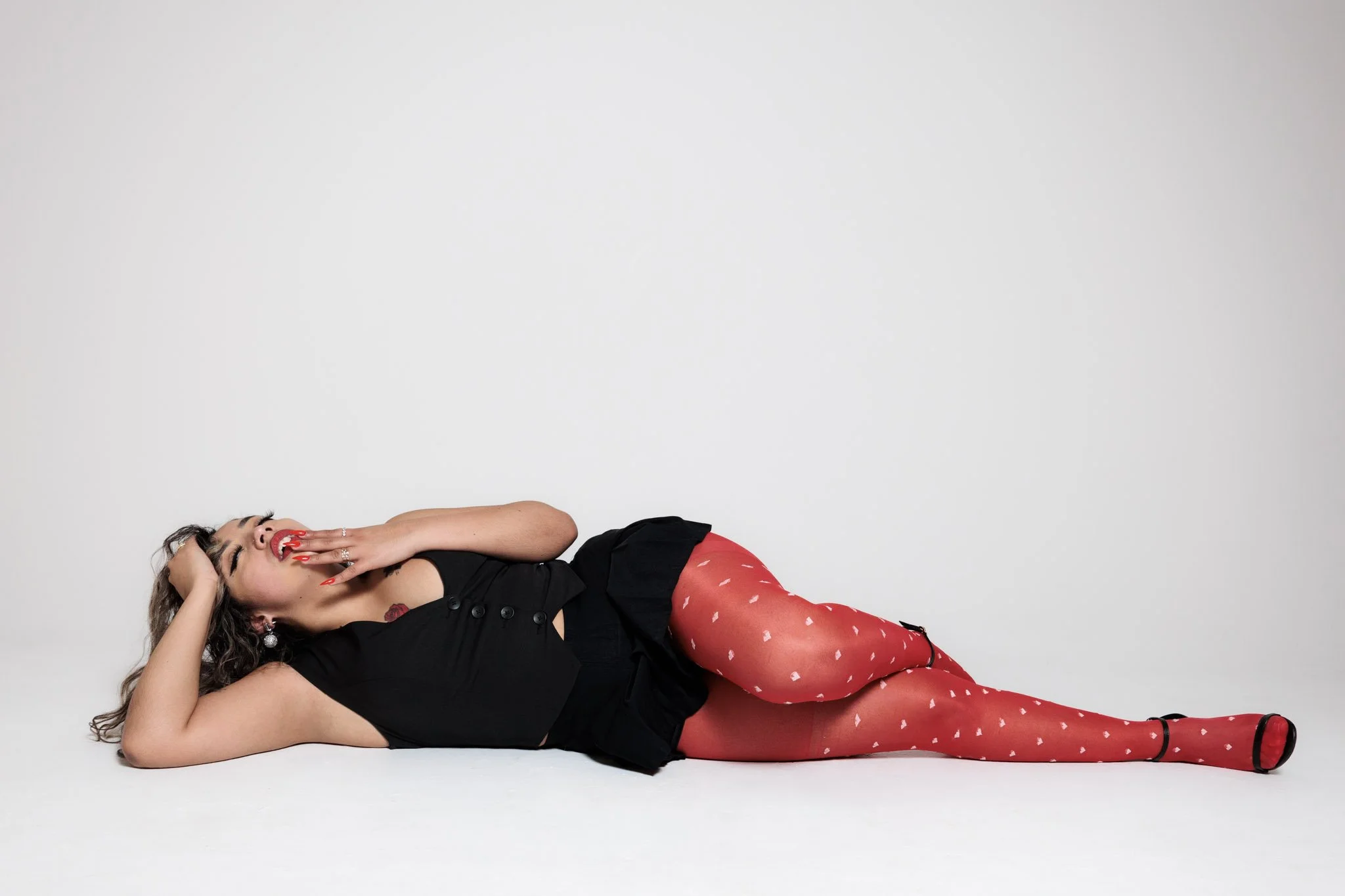 A woman lying on her side on a white surface, wearing red heart-patterned tights, a black dress, and red lipstick, smiling with her hand near her mouth.
