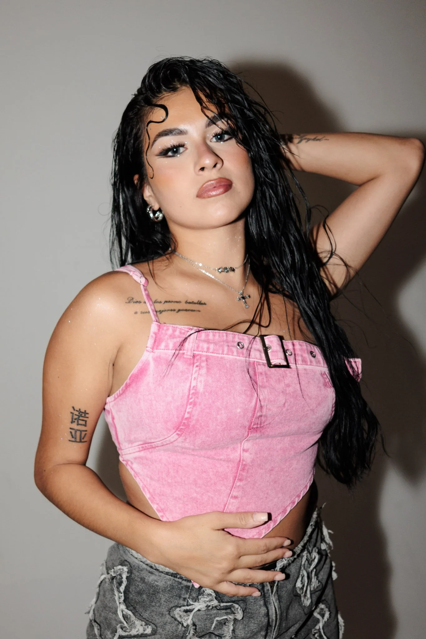 A young woman with long, black, wet hair standing against a plain wall. She is wearing a pink sleeveless top and gray distressed shorts. She has multiple tattoos, jewelry including earrings, necklaces, and rings, and is posing with one hand behind he