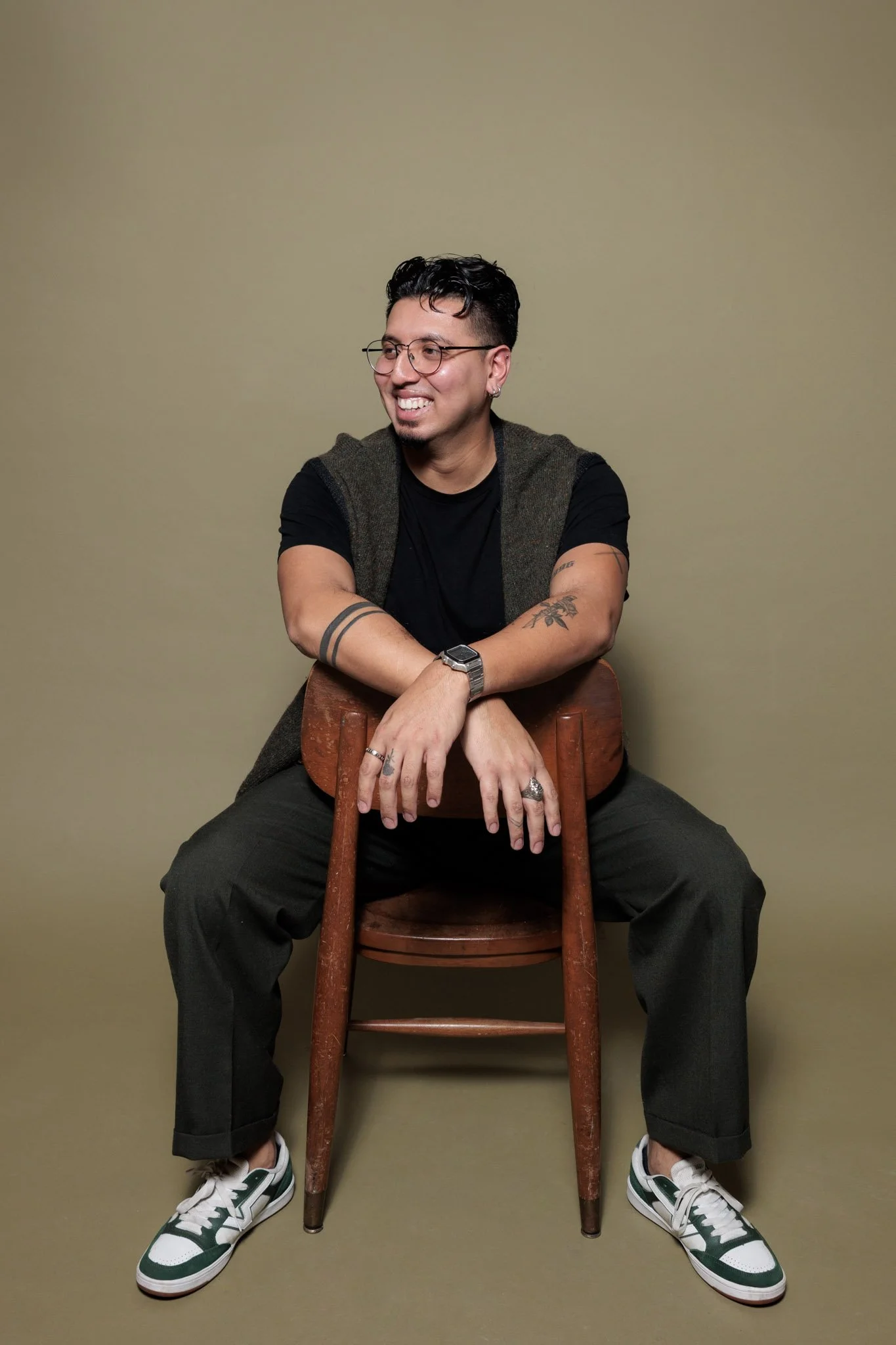 A man sitting on a wooden chair, smiling, wearing glasses, a black T-shirt, black pants, and sneakers, with tattoos and accessories, against a plain beige background.