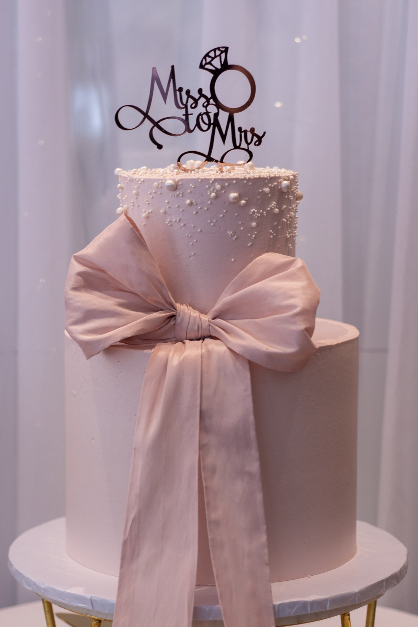 A two-tiered wedding cake decorated with a pink ribbon bow and small white pearl-like accents, topped with a "Mr & Mrs" cake topper featuring a ring and diamond.
