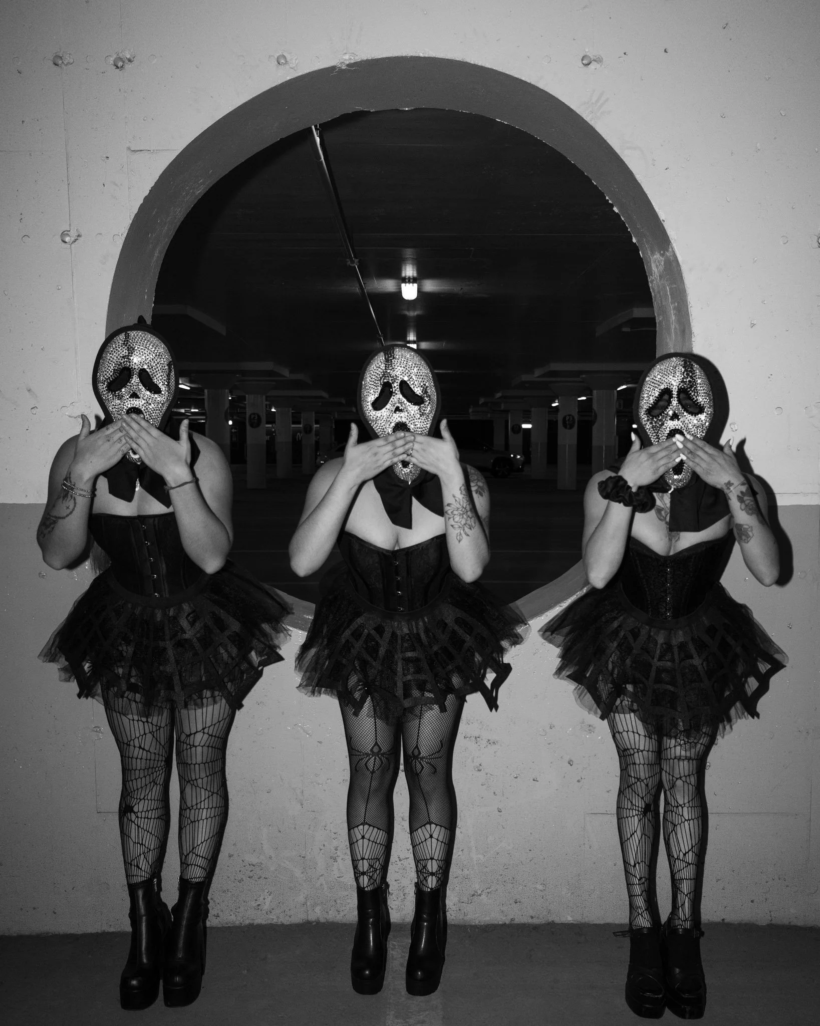 Three women wearing Halloween masks and black costumes, standing near an archway in a parking garage.