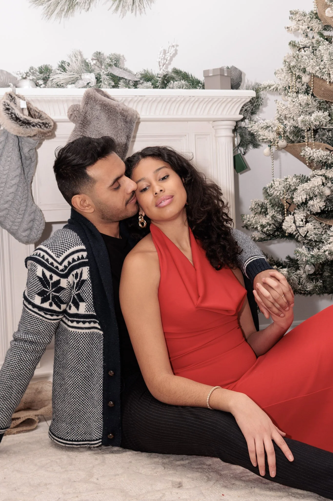 A couple sitting on the floor in front of a Christmas mantel and decorated Christmas tree, with the man leaning in to kiss the woman on the cheek. couple holidays photo session. 