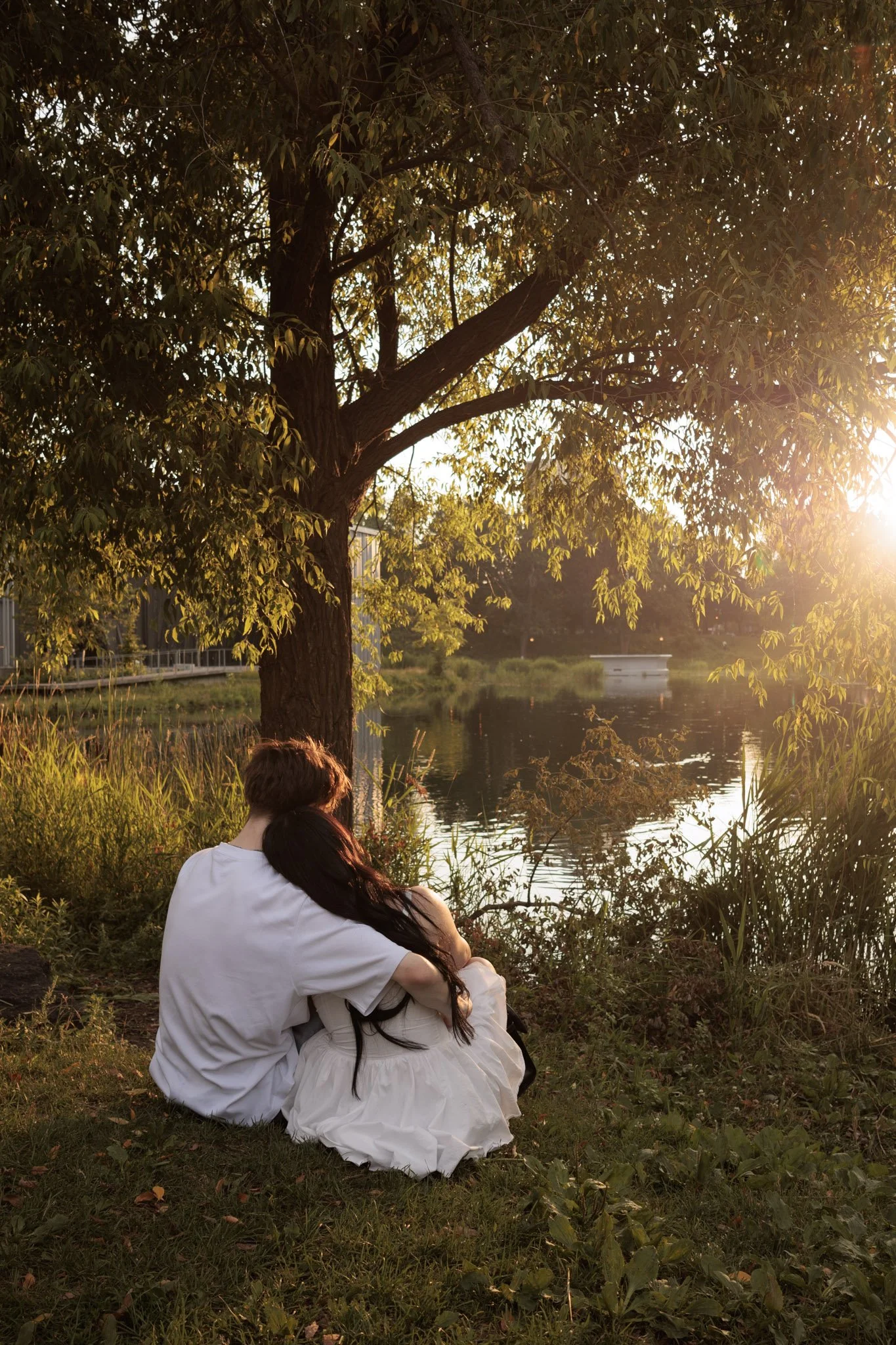 A couple sitting under a large tree by a lake at sunset, with the woman resting her head on the man's shoulder.