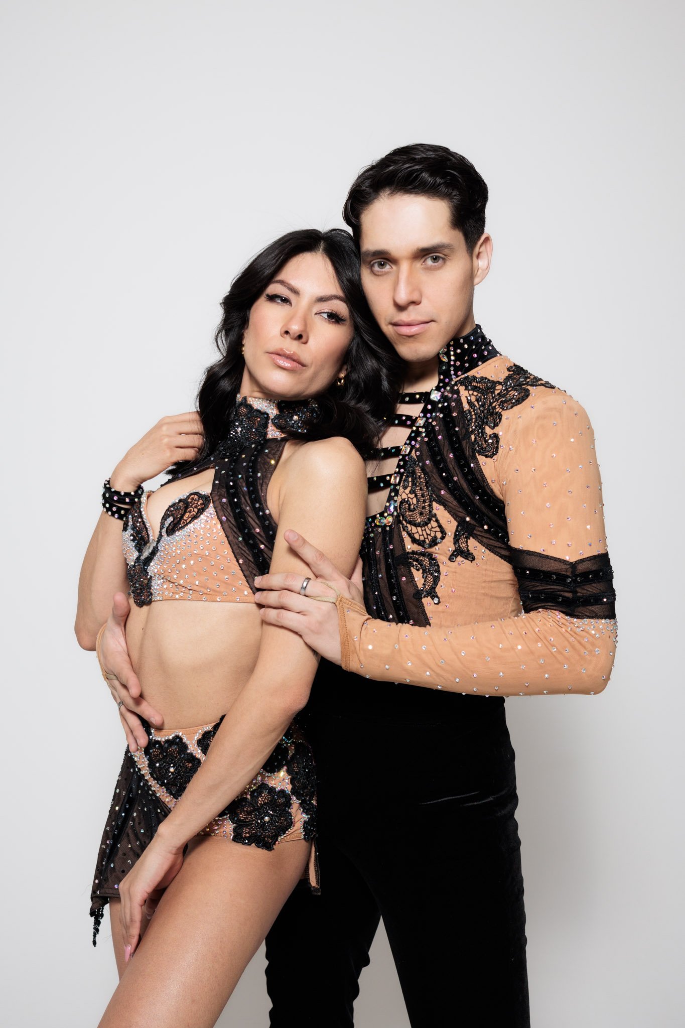 A man and woman posing together against a plain white background, dressed in matching black and peach outfits with lace and rhinestones, with the woman resting her head on the man's shoulder.