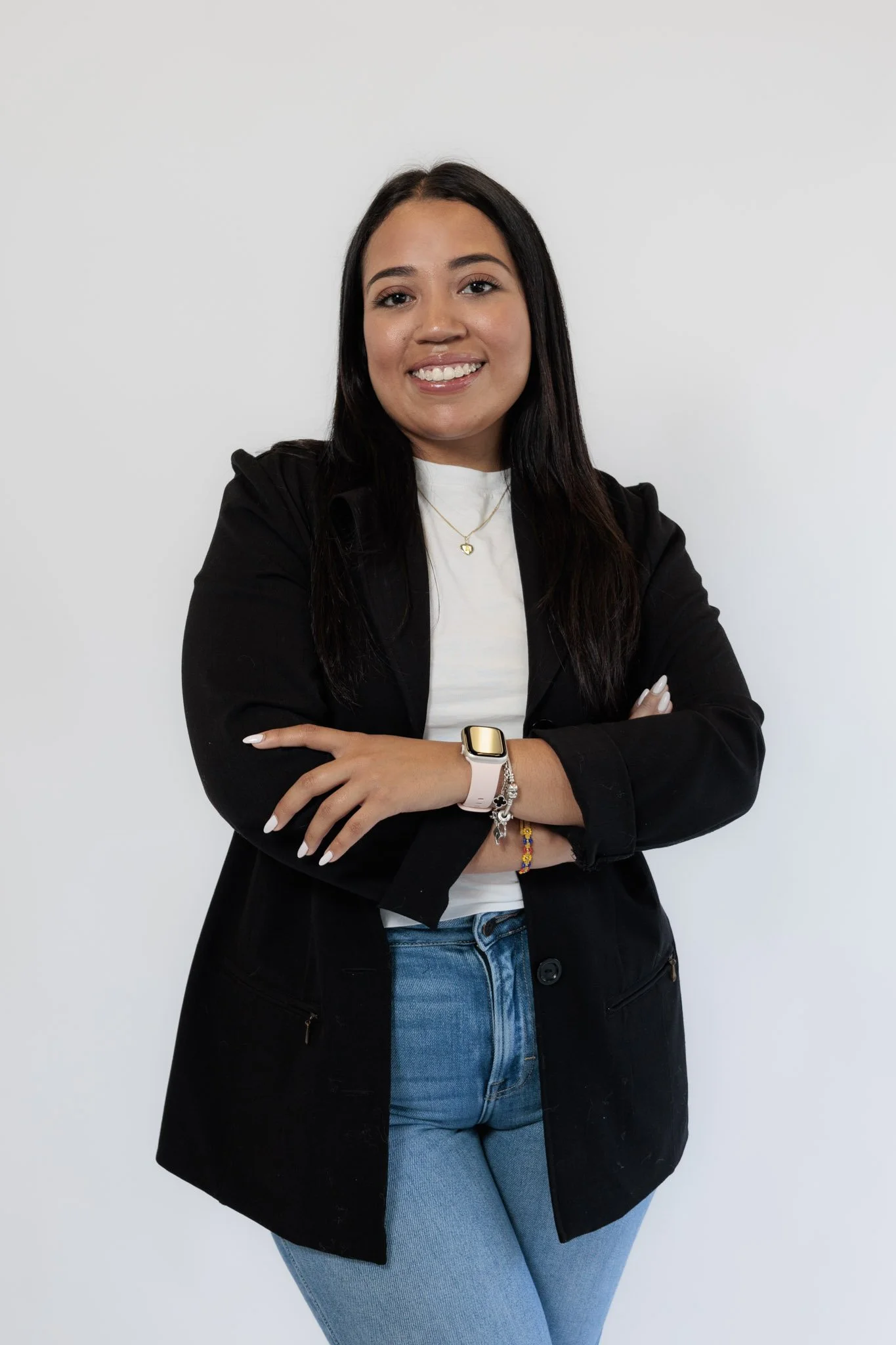 A young woman with long dark hair, wearing a black blazer, white t-shirt, and blue jeans, standing with arms crossed against a plain white background.