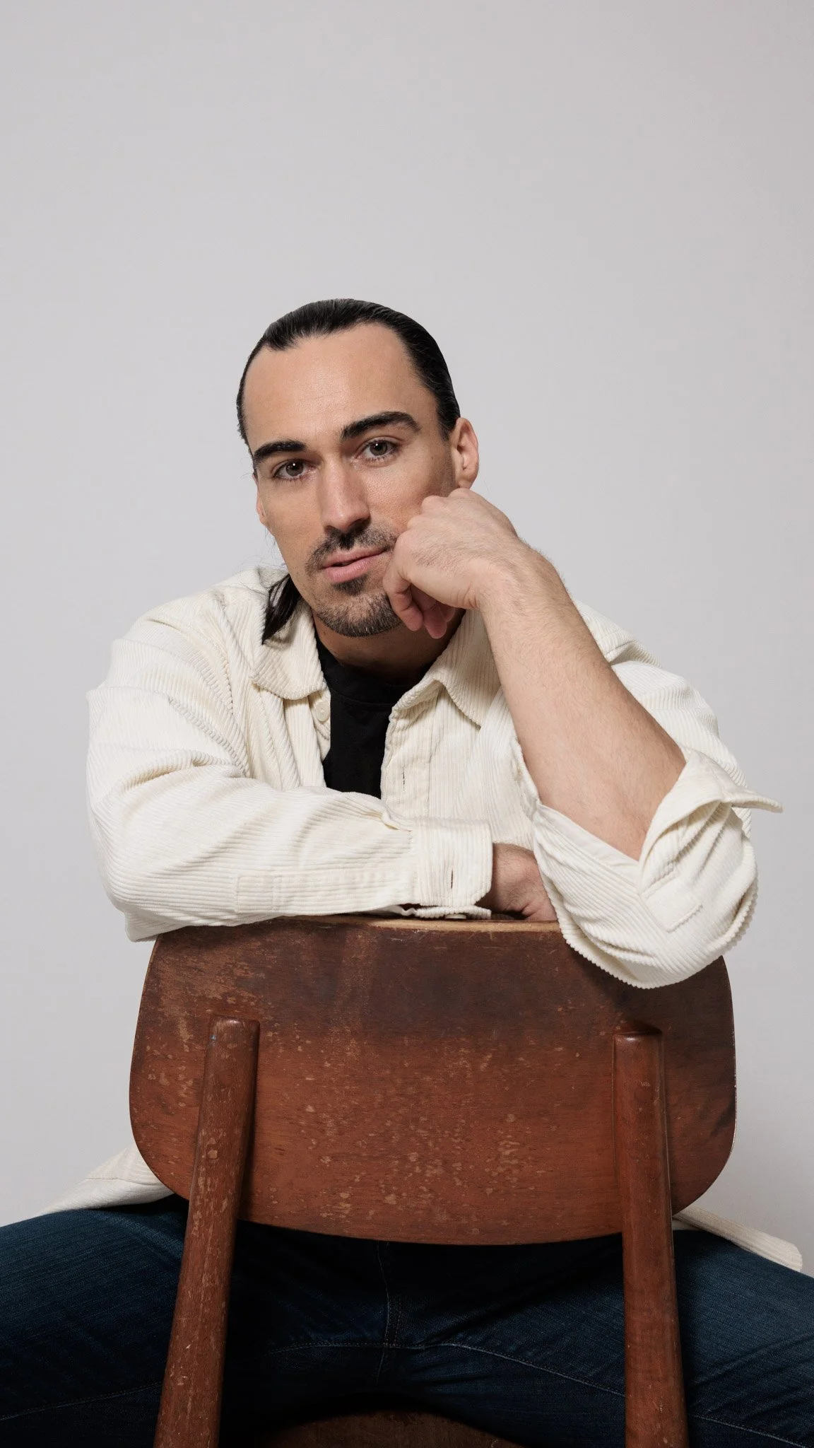 Portrait of a man sitting behind a wooden chair, resting his chin on his hand, against a plain white background.