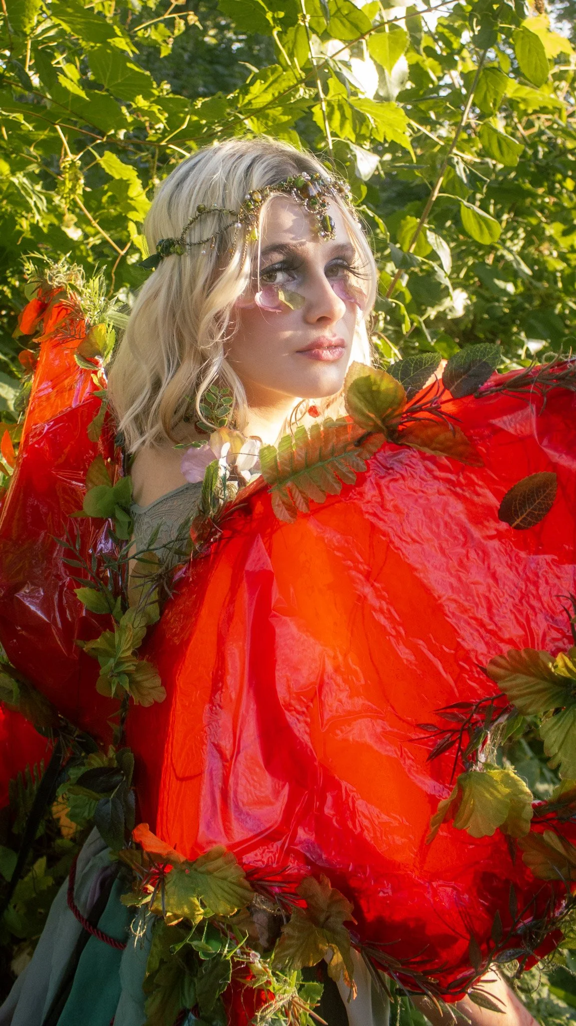 A woman with blonde hair dressed in an elaborate costume made of leaves and red plastic, standing outdoors in a green, leafy environment.