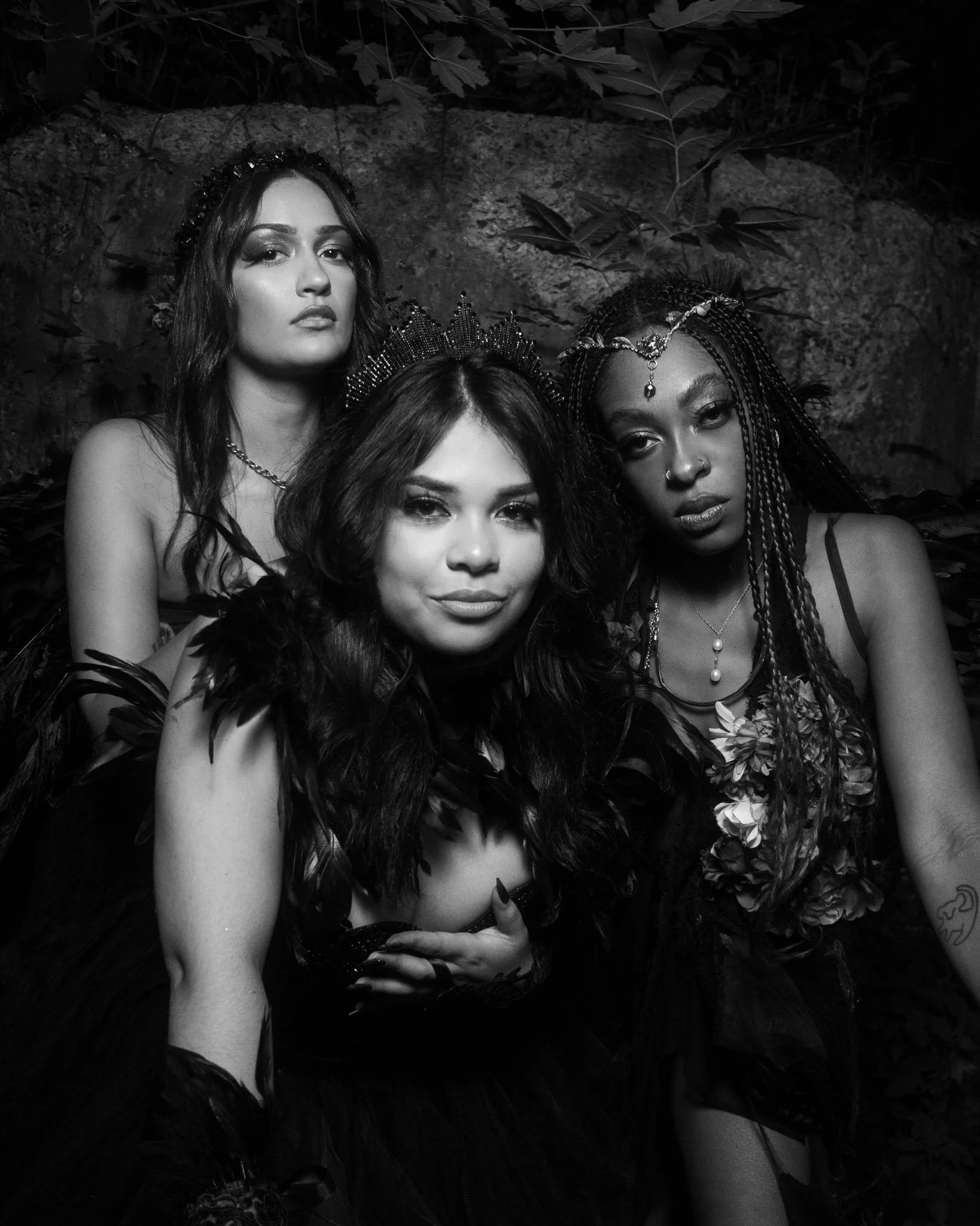 Three women dressed in ornate costumes with crowns and jewelry, posing together. One woman has long, dark hair and a crown, the second has wavy hair and a crown, and the third has braided hair with jewelry on her forehead, all set against a natural b