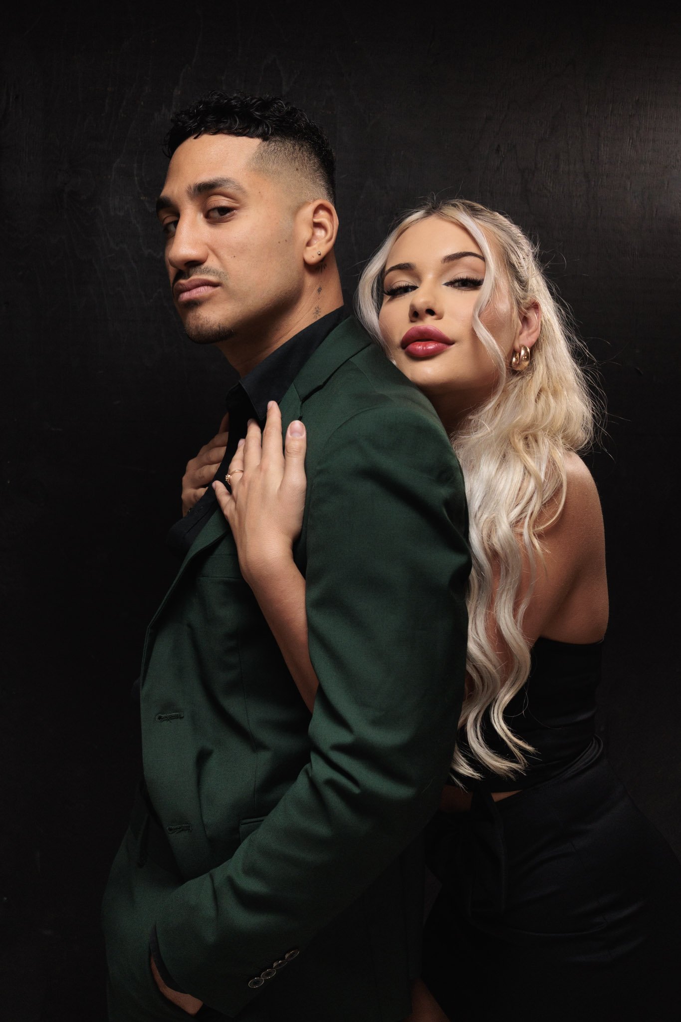A man and woman posing together against a dark background, with the woman standing behind the man, her hand on his shoulder, both looking confidently at the camera. Couple photography. 