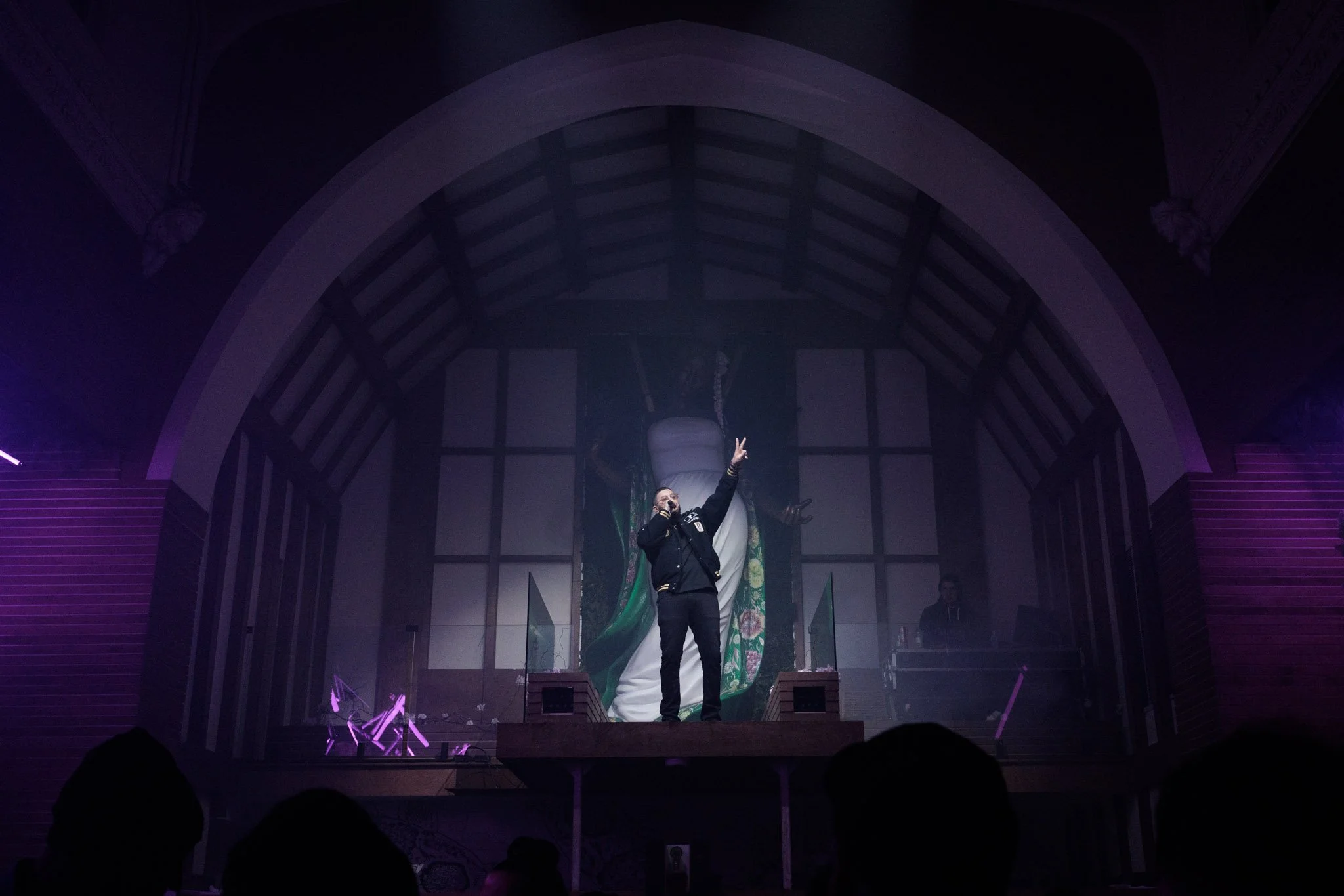 A man standing on a stage with a microphone, gesturing upwards with his right hand, in front of a large backdrop featuring a giant ceramic vase. The stage is inside a room with arched ceilings and dim purple-purple lighting, with an audience silhouet