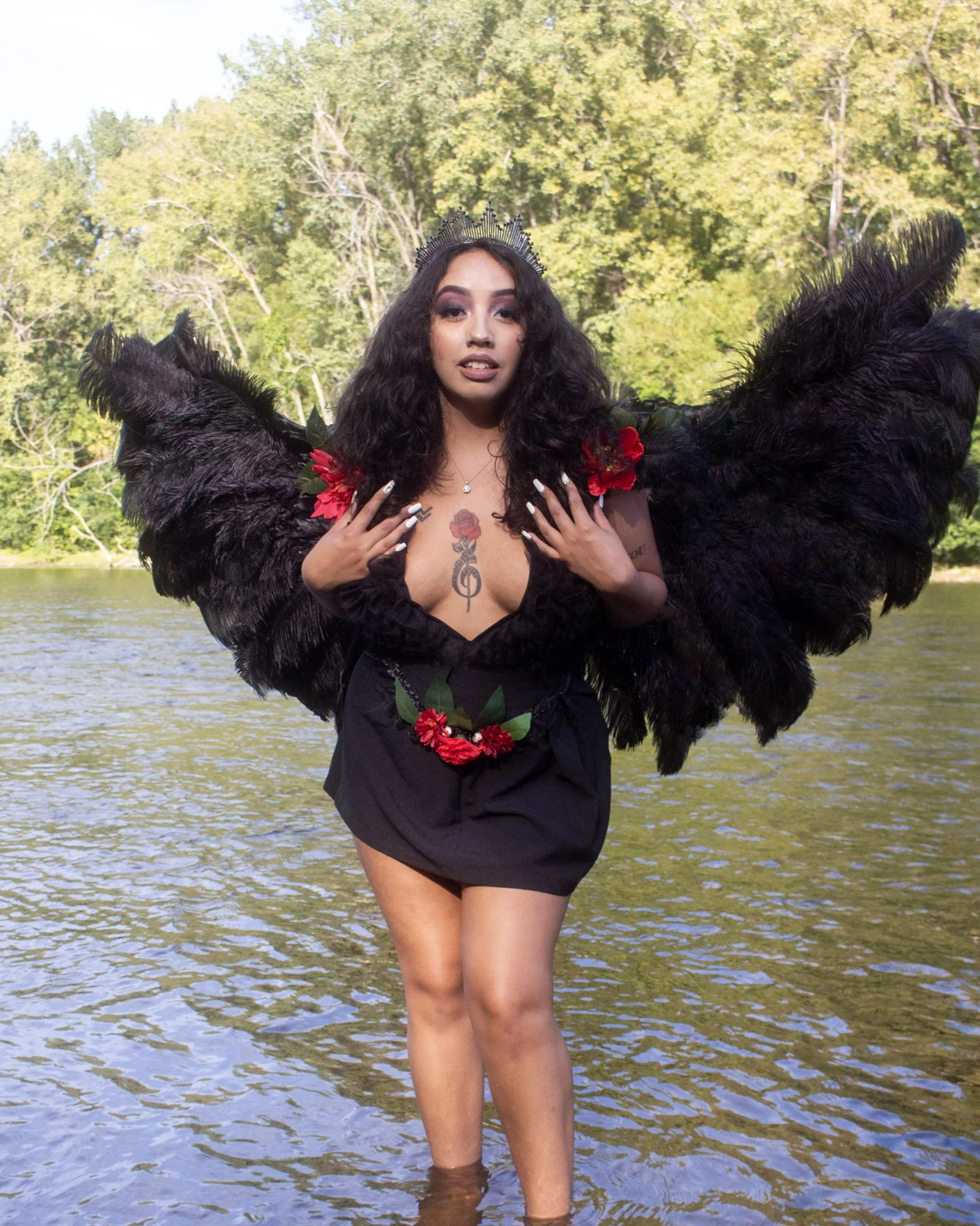 A woman standing in water near trees, wearing a black dress with large black feathered wings, a crown, and red flowers on her shoulders and waist, with tattoos visible on her chest and arm.