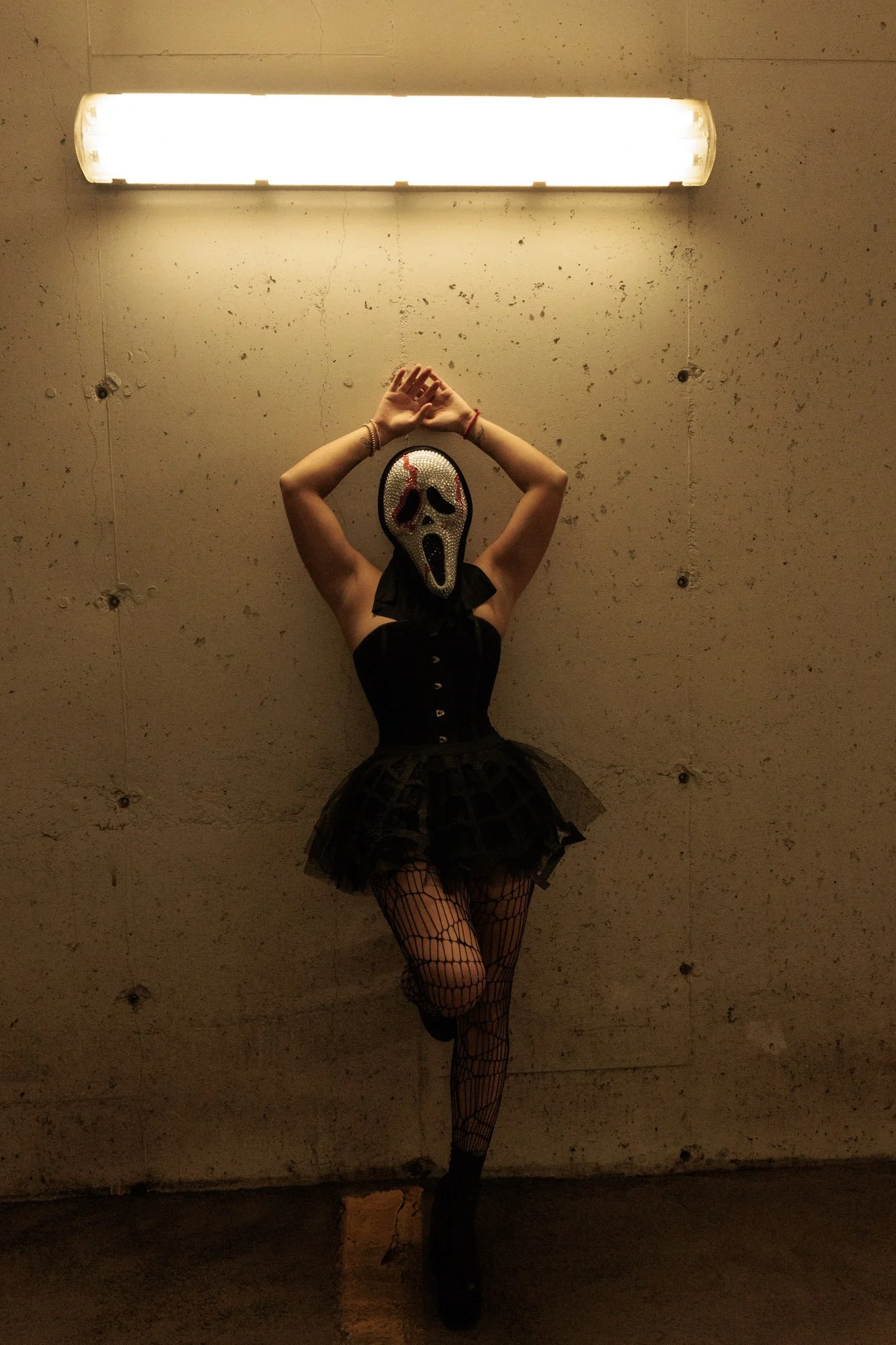 Person wearing a mask resembling the scream mask from the movie Scream, dressed in a black dress and fishnet stockings, standing against a concrete wall with a bright light above.